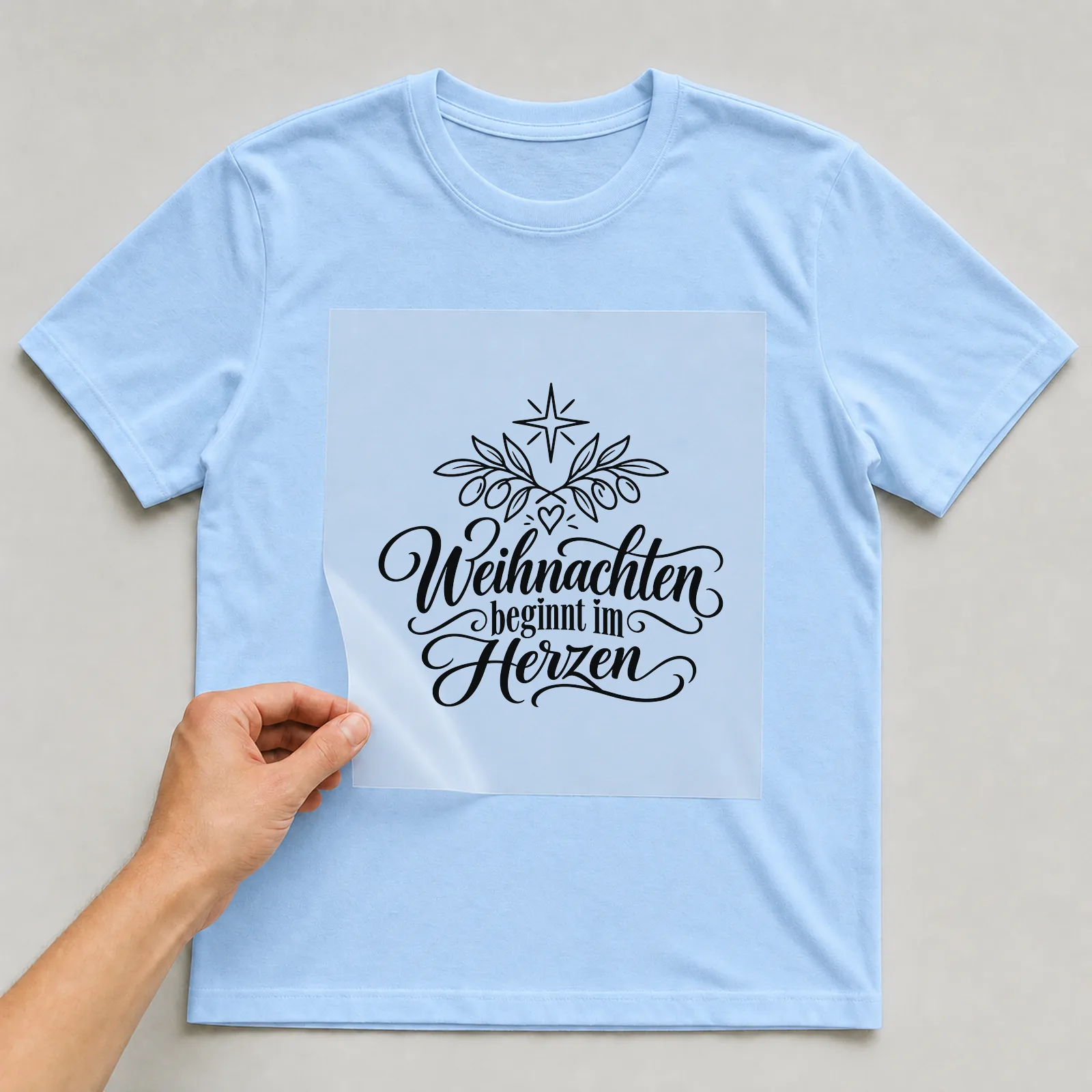 Bügelbild „Weihnachten beginnt im Herzen“ auf hellblauem T-Shirt, Folie wird aufgelegt