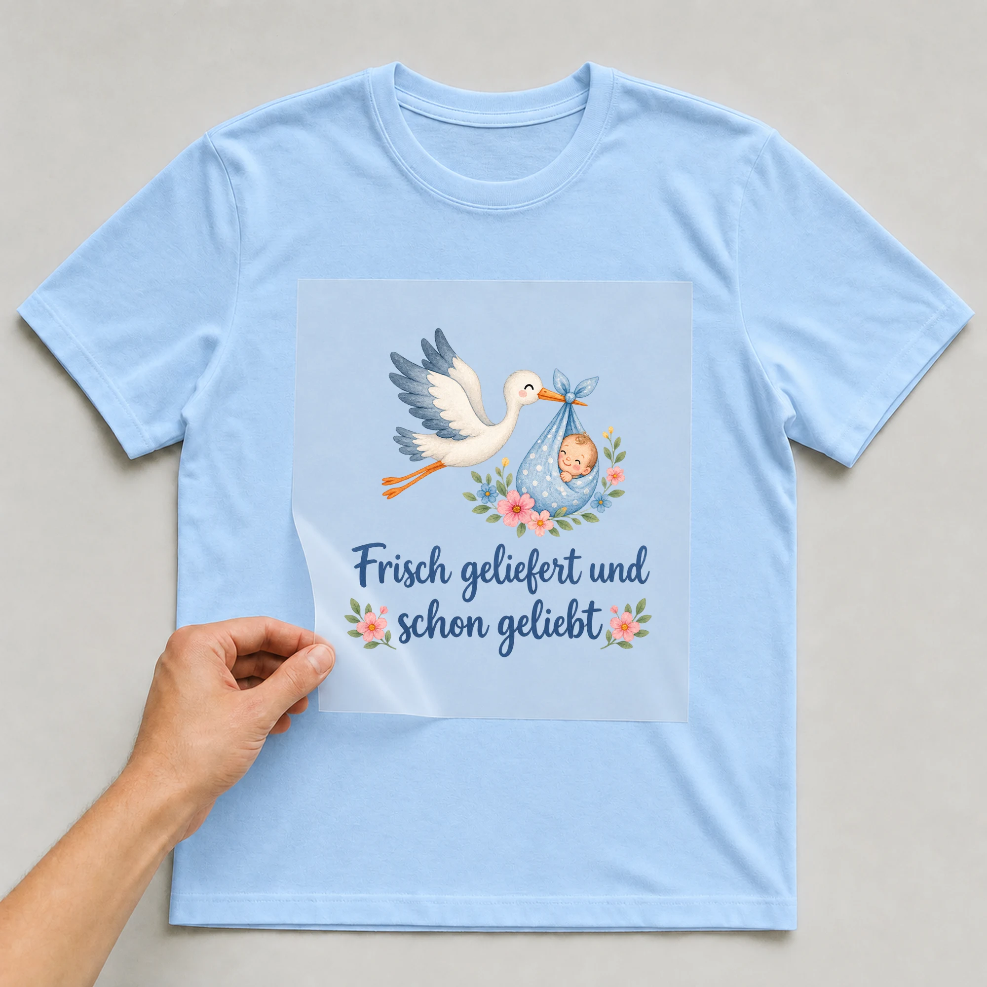 Bügelbild „Frisch geliefert und schon geliebt“ auf babyblauem T-Shirt, Folie wird aufgelegt
