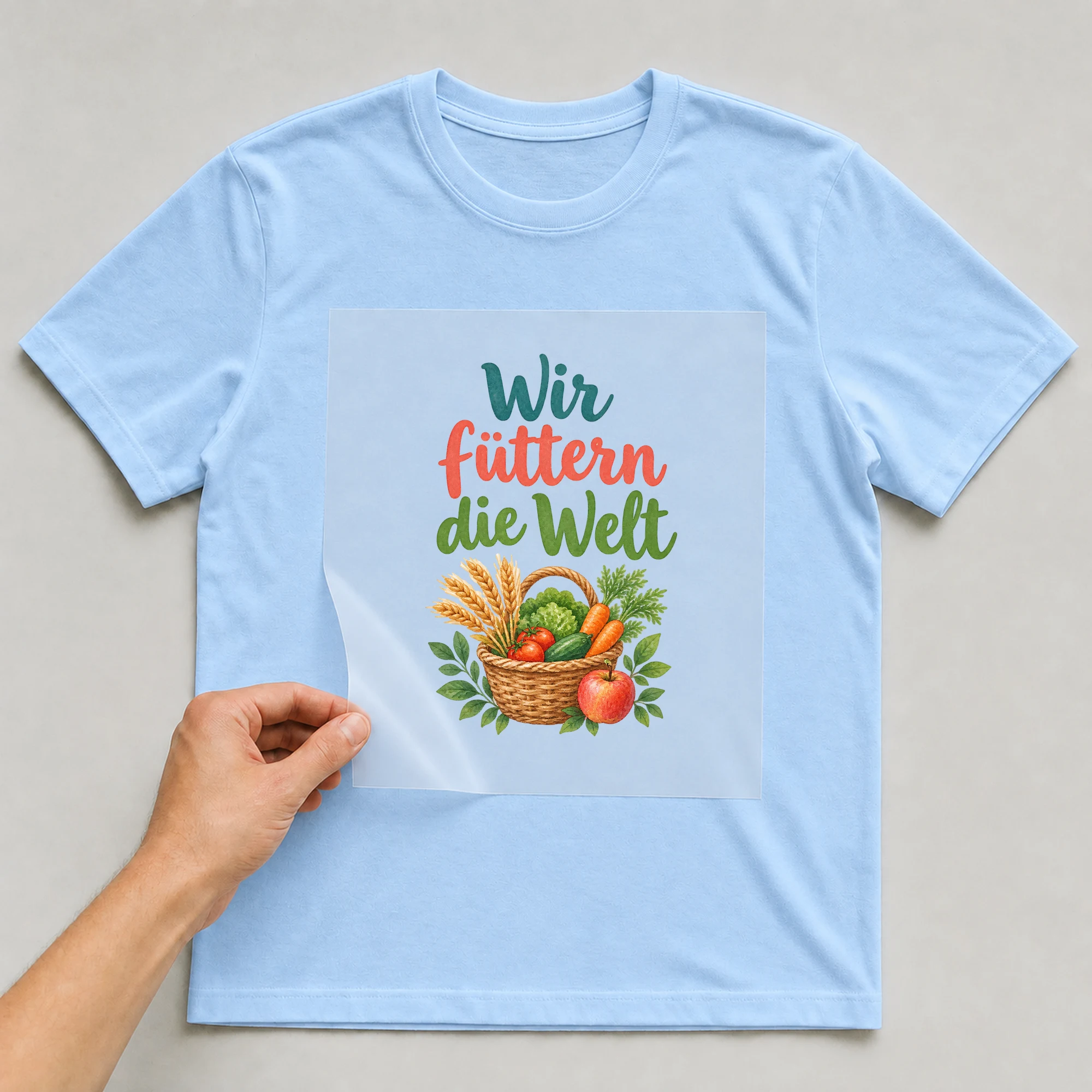 Bügelbild „Wir füttern die Welt“ auf babyblauem T-Shirt, Folie wird aufgelegt