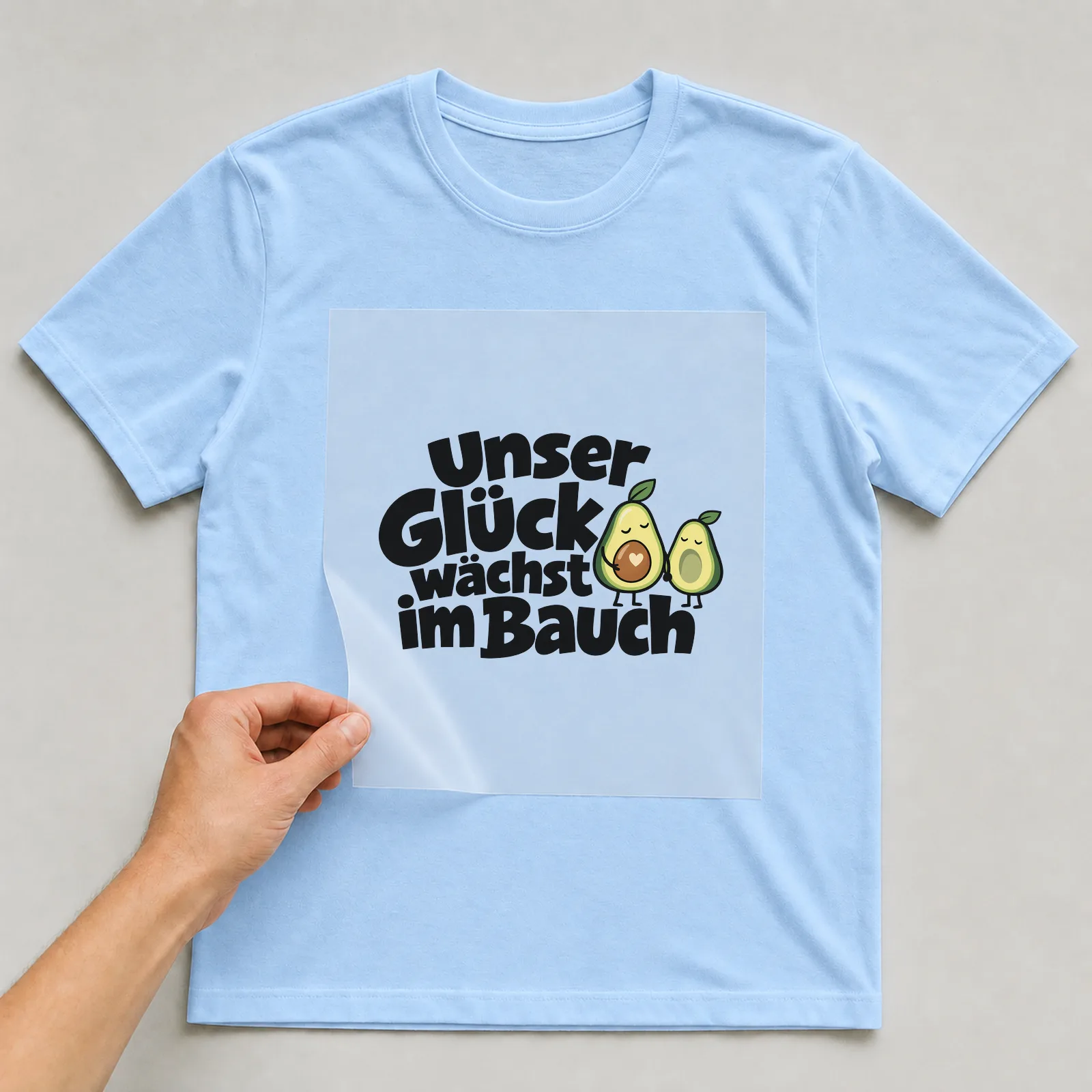 Bügelbild „Unser Glück wächst im Bauch“ auf hellblauem T-Shirt, Folie wird aufgelegt
