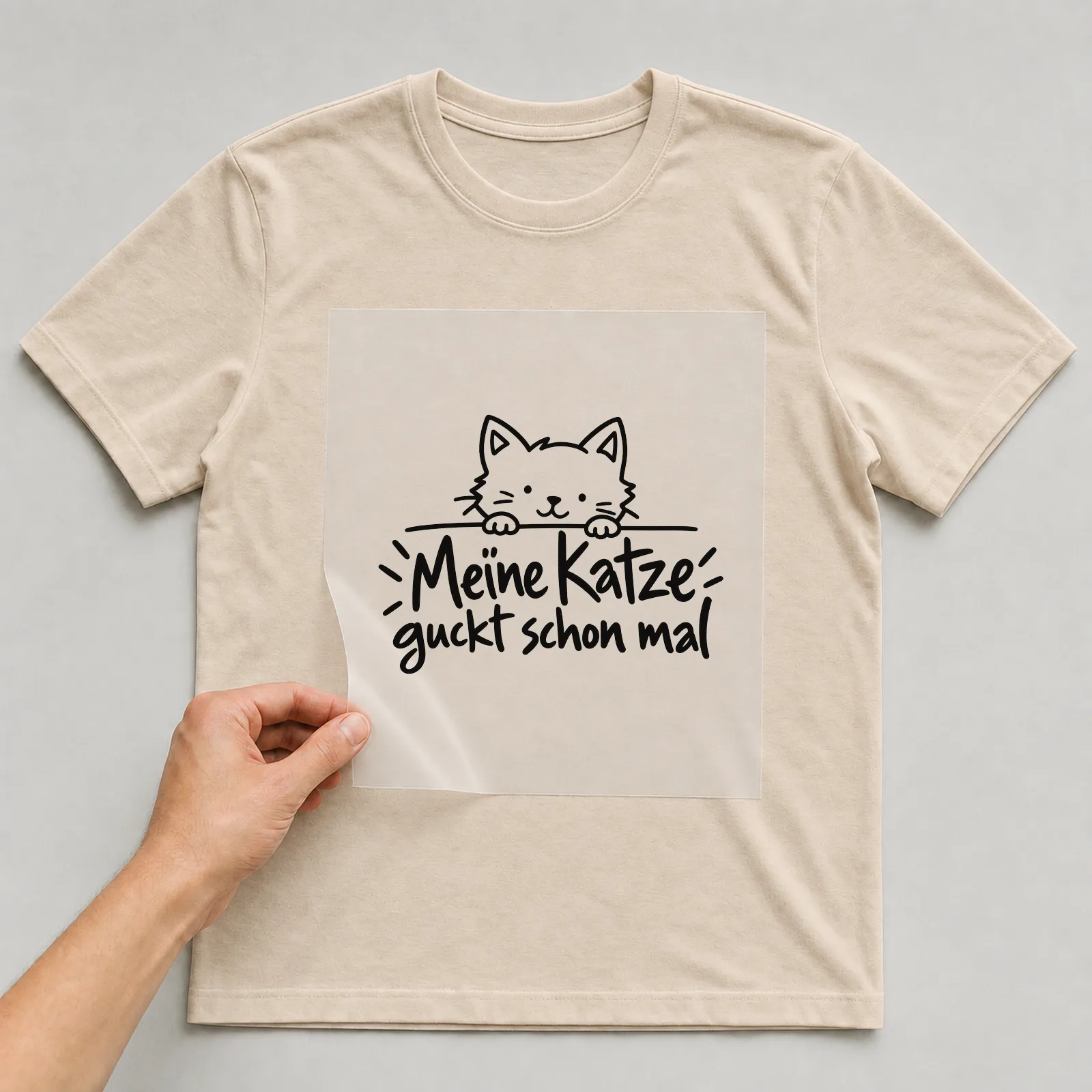 Bügelbild „Meine Katze guckt schon mal“ auf beigem T-Shirt, Folie wird aufgelegt