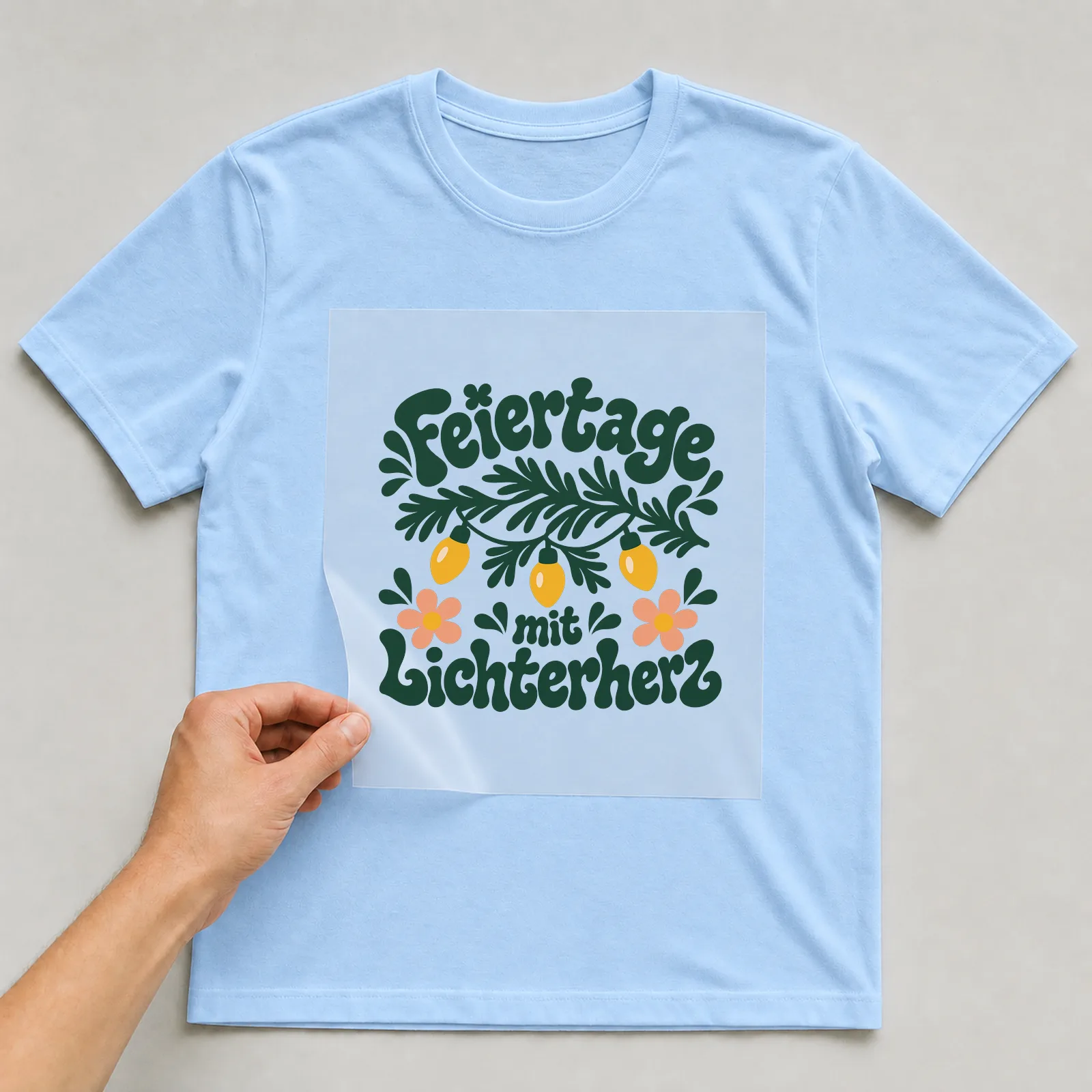 Bügelbild „Feiertage mit Lichterherz“ auf hellblauem T-Shirt, Folie wird aufgelegt