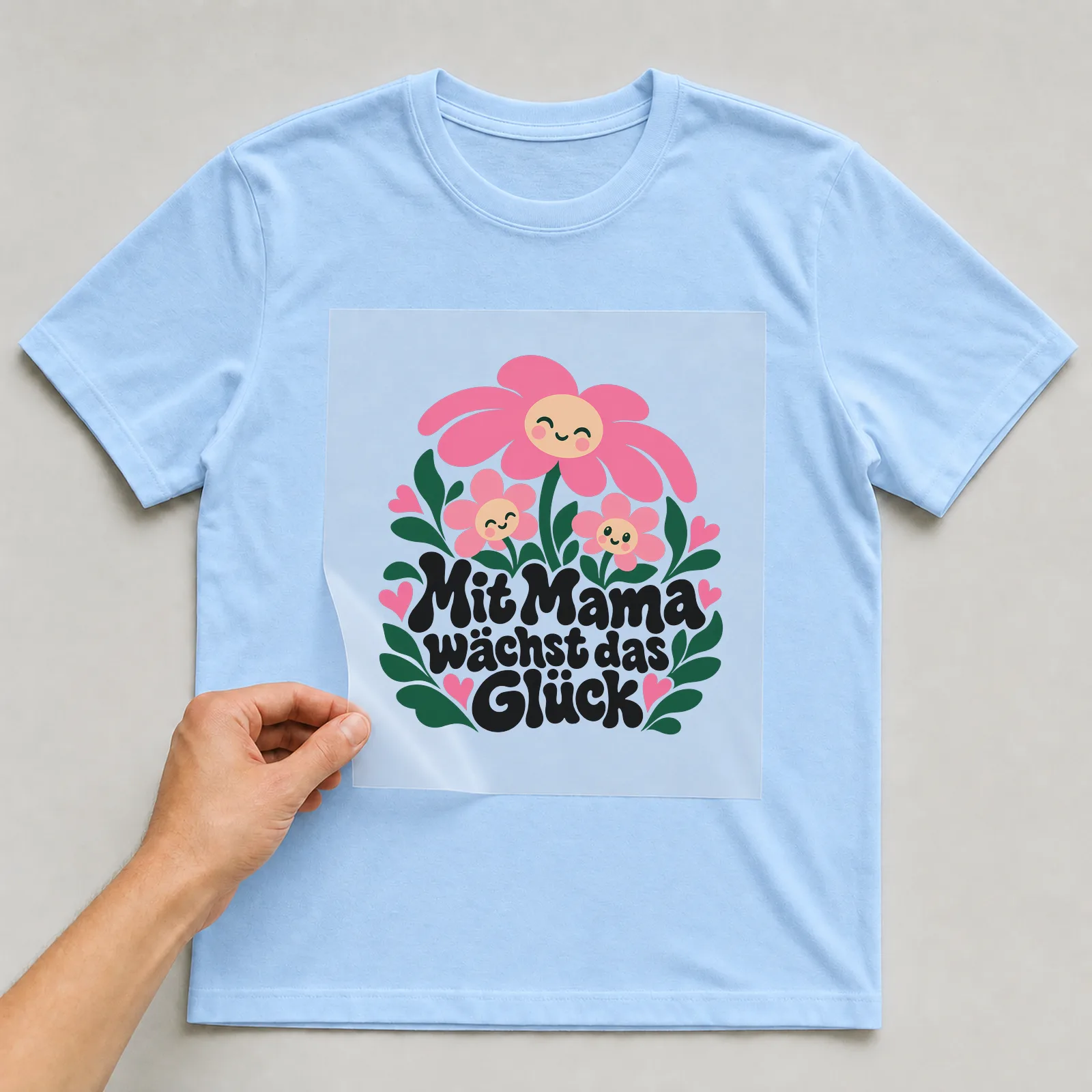 Bügelbild „Mit Mama wächst das Glück“ auf hellblauem T-Shirt, Folie wird aufgelegt