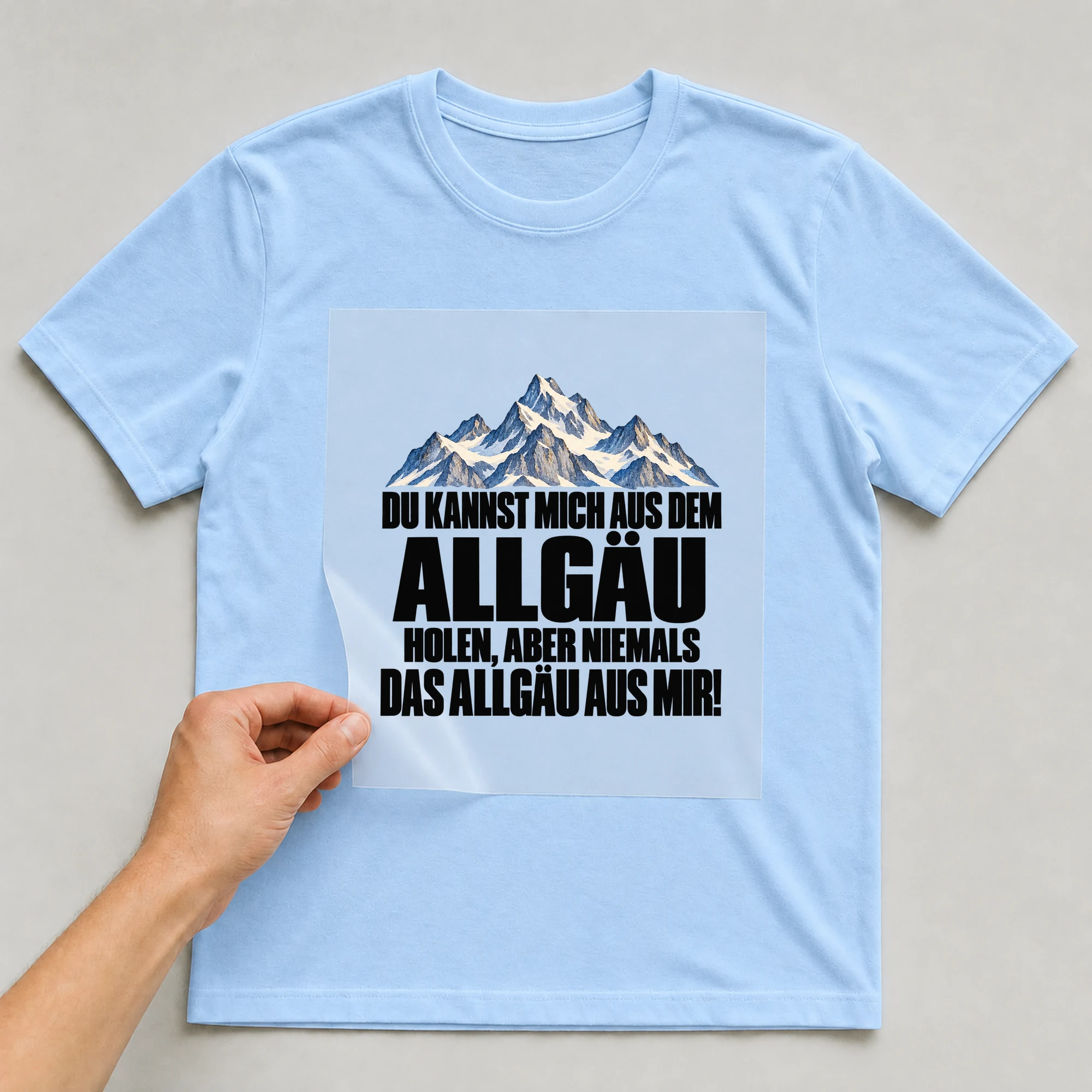 Bügelbild „Du Kannst Mich Aus Dem Allgäu Holen, Aber Niemals Das Allgäu Aus Mir!“ auf babyblauem T-Shirt, Folie wird aufgelegt