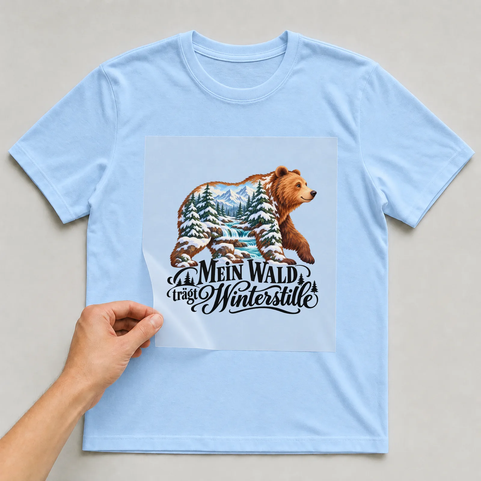 Bügelbild „Mein Wald trägt Winterstille“ auf hellblauem T-Shirt, Folie wird aufgelegt