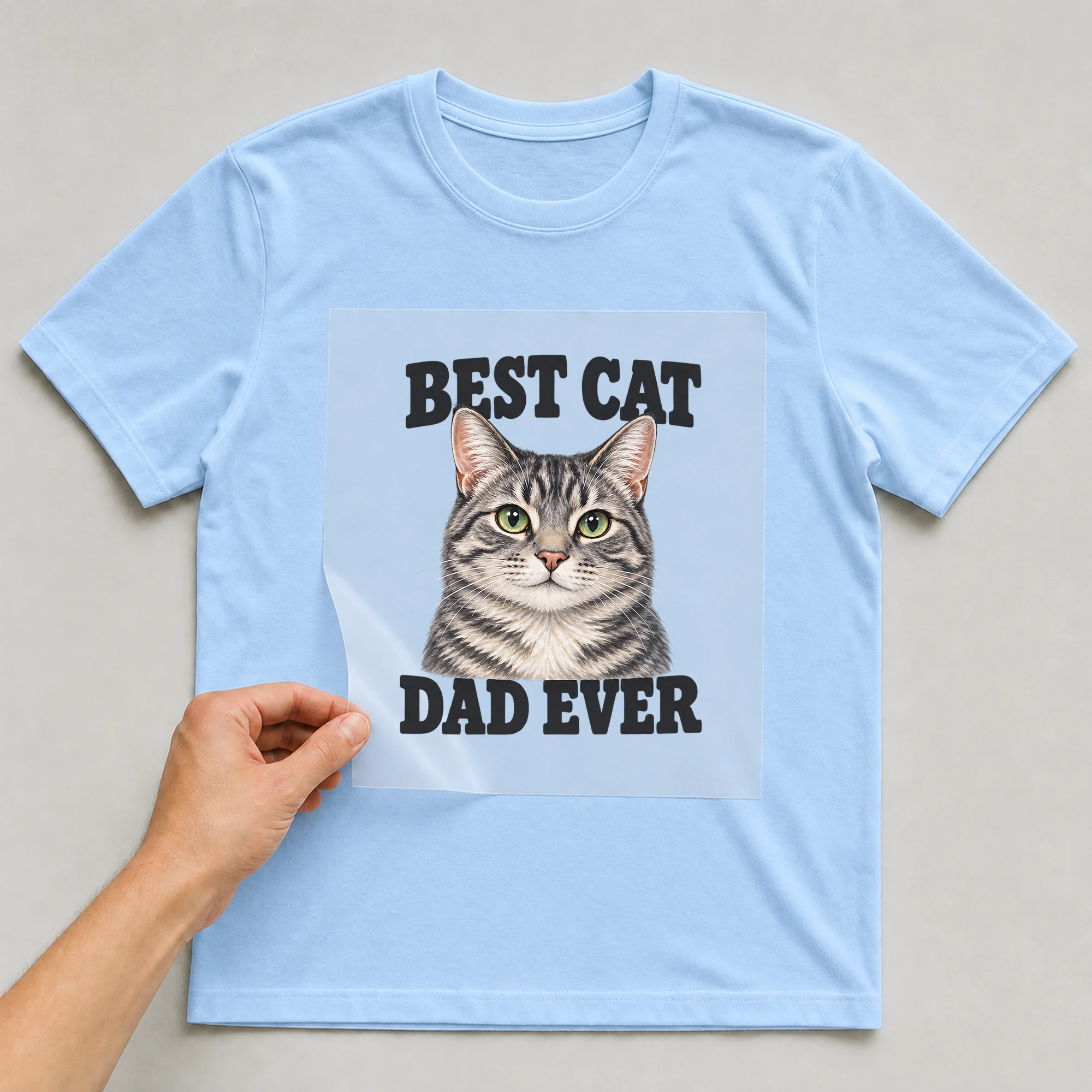 Bügelbild „Best Cat Dad Ever“ auf babyblauem T-Shirt, Folie wird aufgelegt