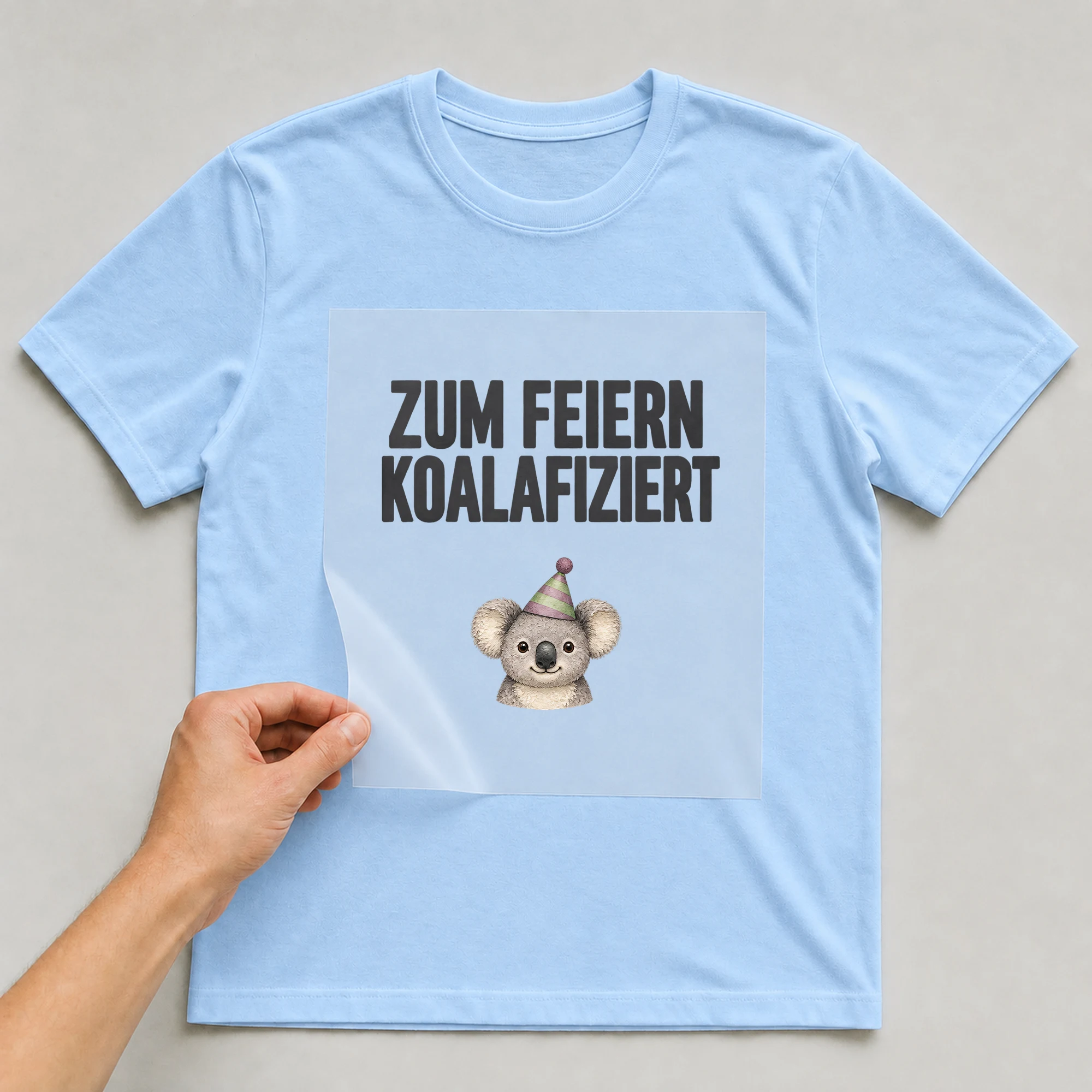 Bügelbild „ZUM FEIERN KOALAFIZIERT“ auf babyblauem T-Shirt, Folie wird aufgelegt