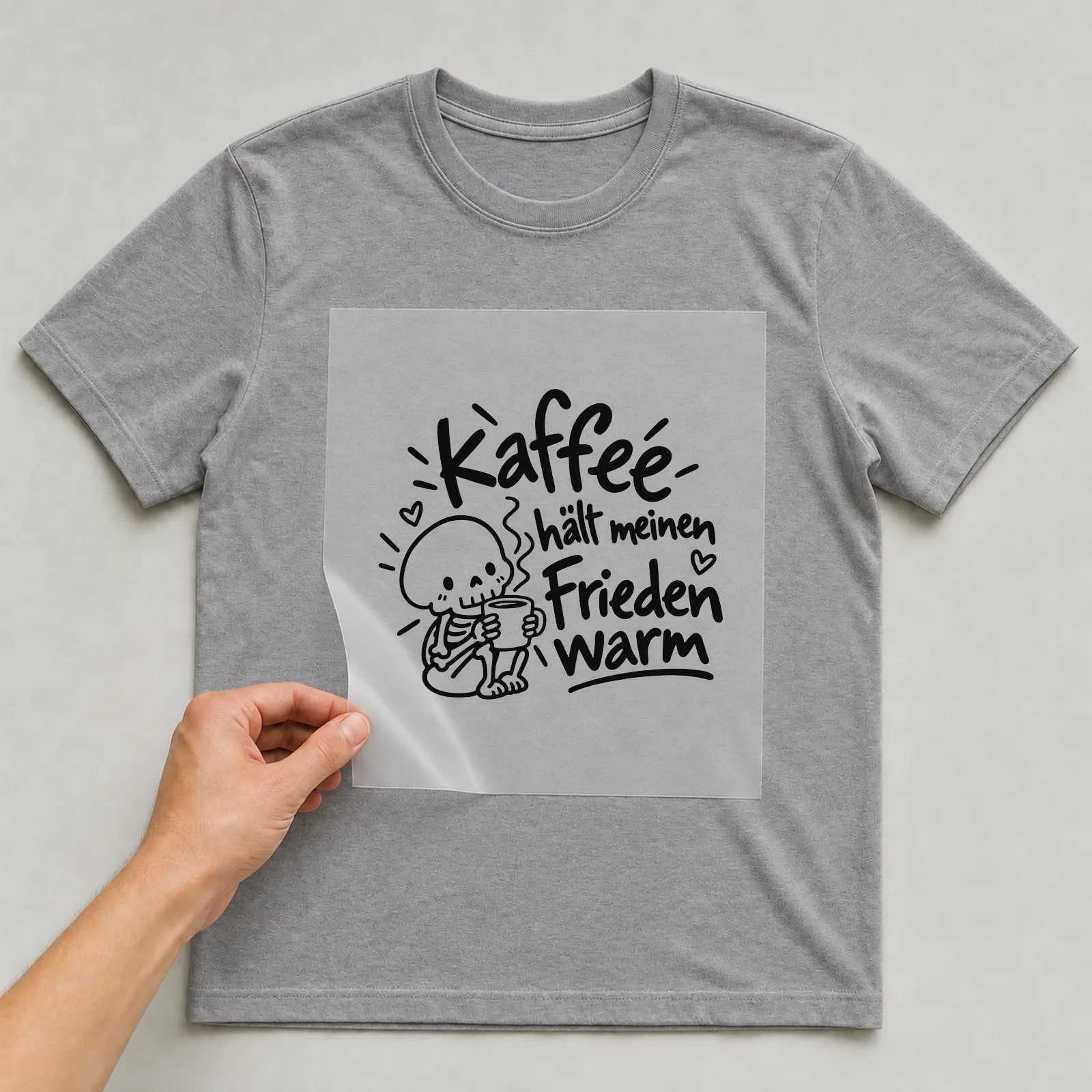 Bügelbild „Kaffee hält meinen Frieden warm“ auf grauem T-Shirt, Folie wird aufgelegt