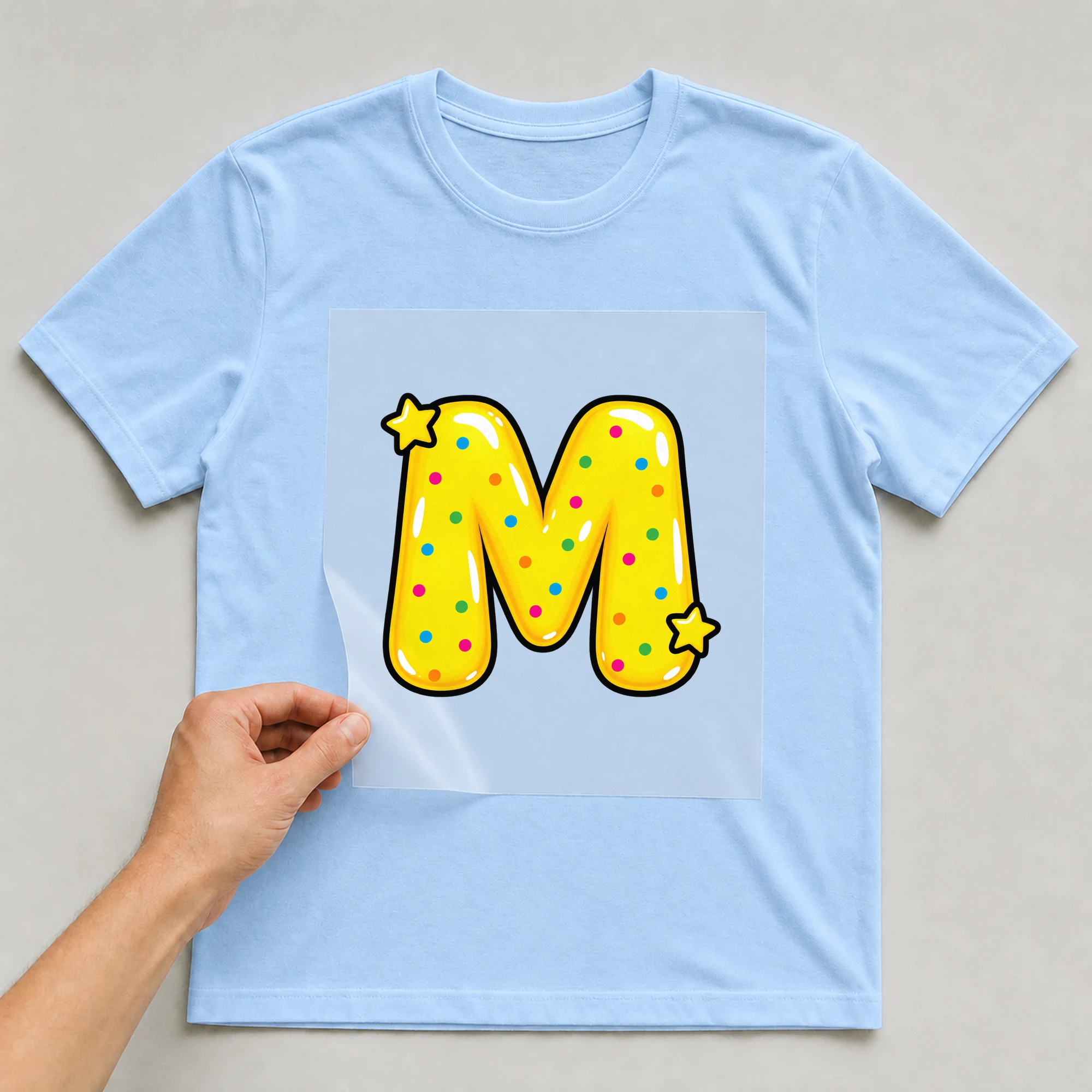 Bügelbild „Buchstabe M im Comic-Ballon-Stil“ auf babyblauem T-Shirt, Folie wird aufgelegt