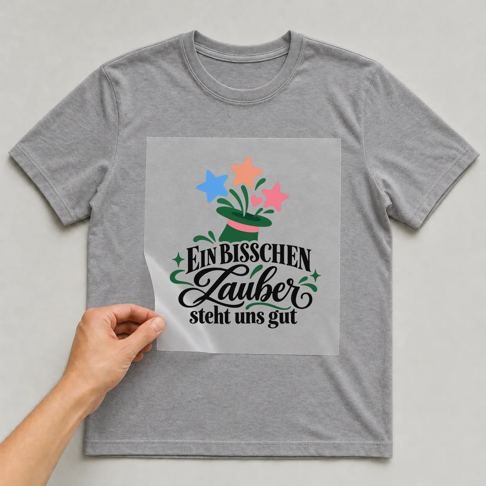 Bügelbild „EIN BISSCHEN Zauber steht uns gut“ auf grauem T-Shirt, Folie wird aufgelegt