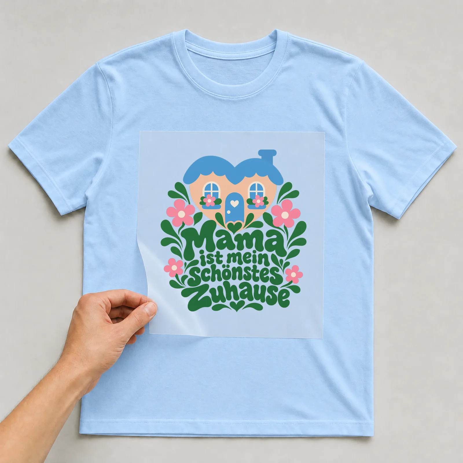 Bügelbild „Mama ist mein schönstes Zuhause“ auf hellblauem T-Shirt, Folie wird aufgelegt