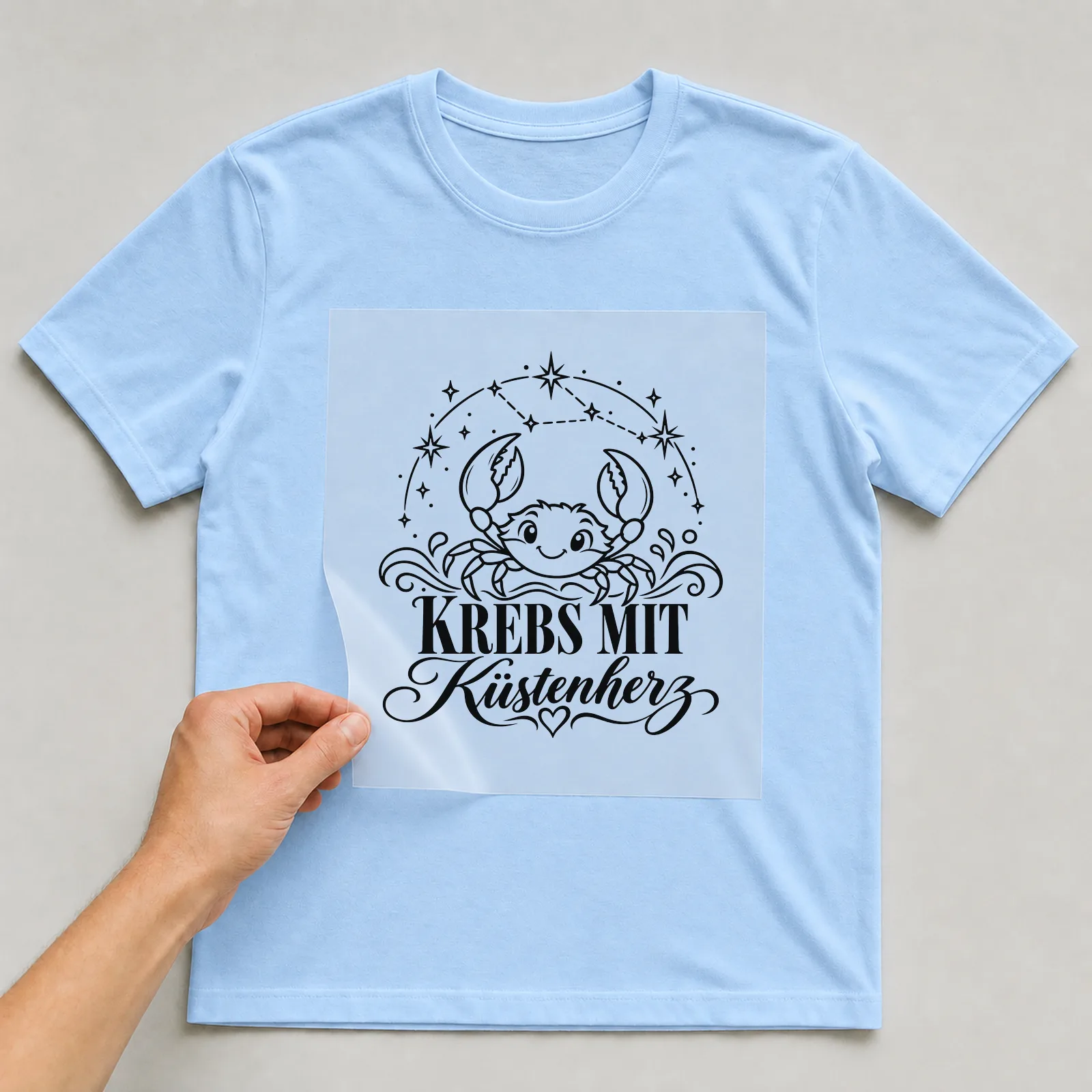 Bügelbild „Krebs mit Küstenherz“ auf hellblauem T-Shirt, Folie wird aufgelegt