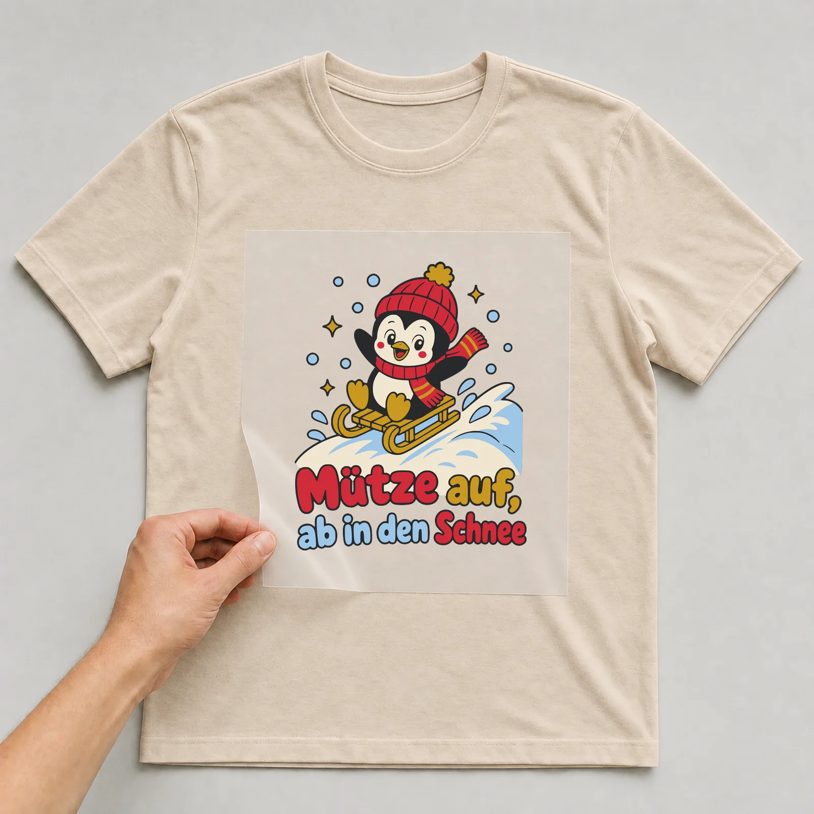 Bügelbild Mütze auf, ab in den Schnee auf beigem T-Shirt, Folie wird aufgelegt