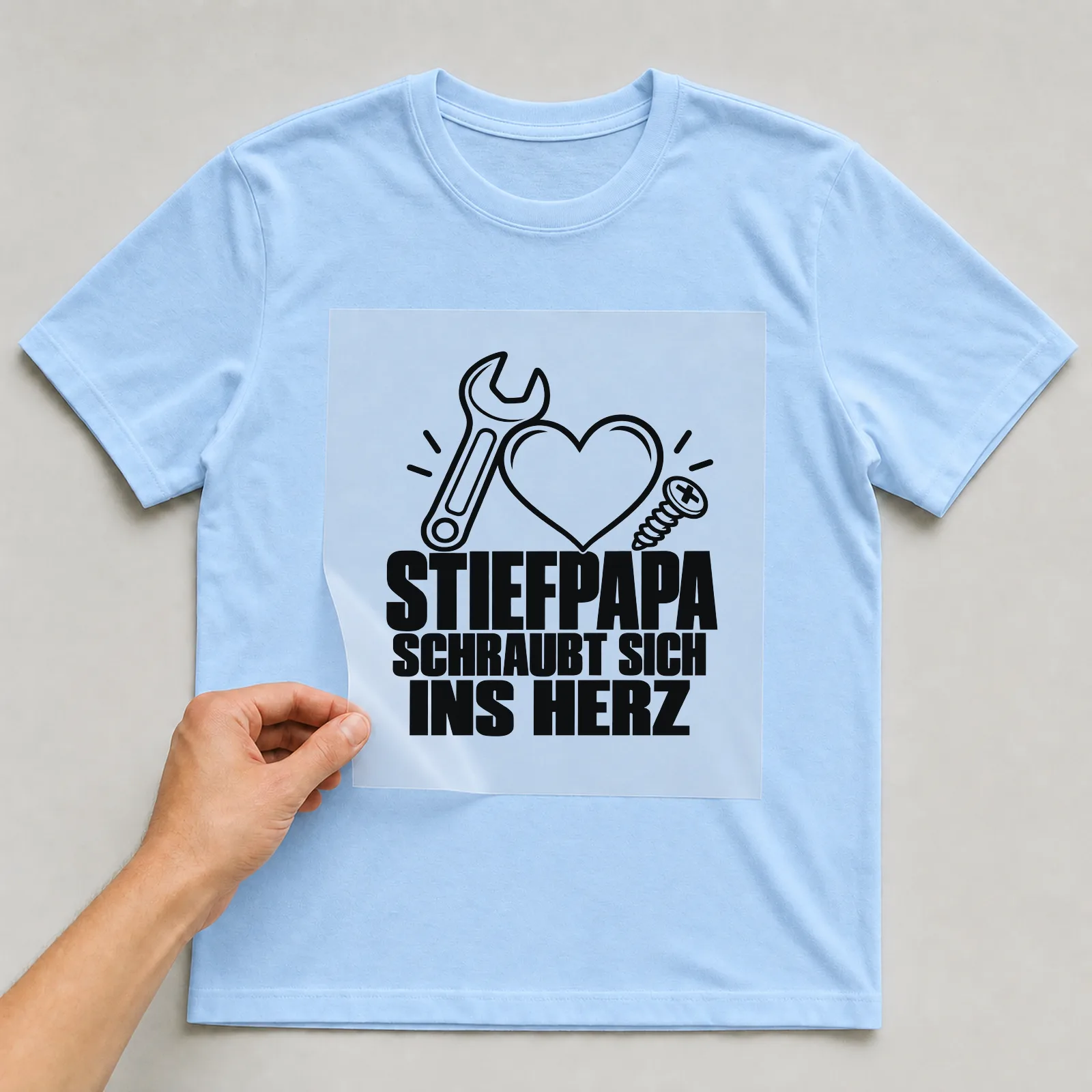 Bügelbild „Stiefpapa schraubt sich ins Herz“ auf hellblauem T-Shirt, Folie wird aufgelegt
