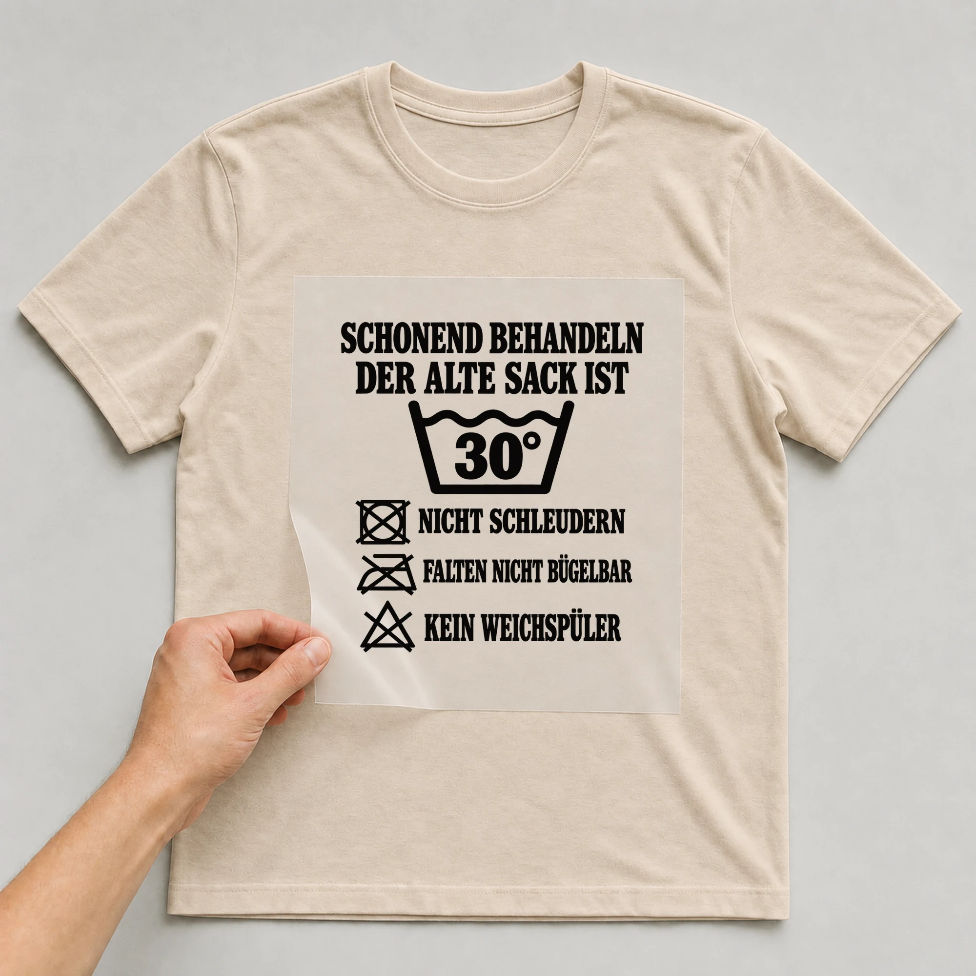 Bügelbild „Schonend behandeln der alte Sack ist 30° nicht schleudern falten nicht“ auf beigem T-Shirt, Folie wird aufgelegt