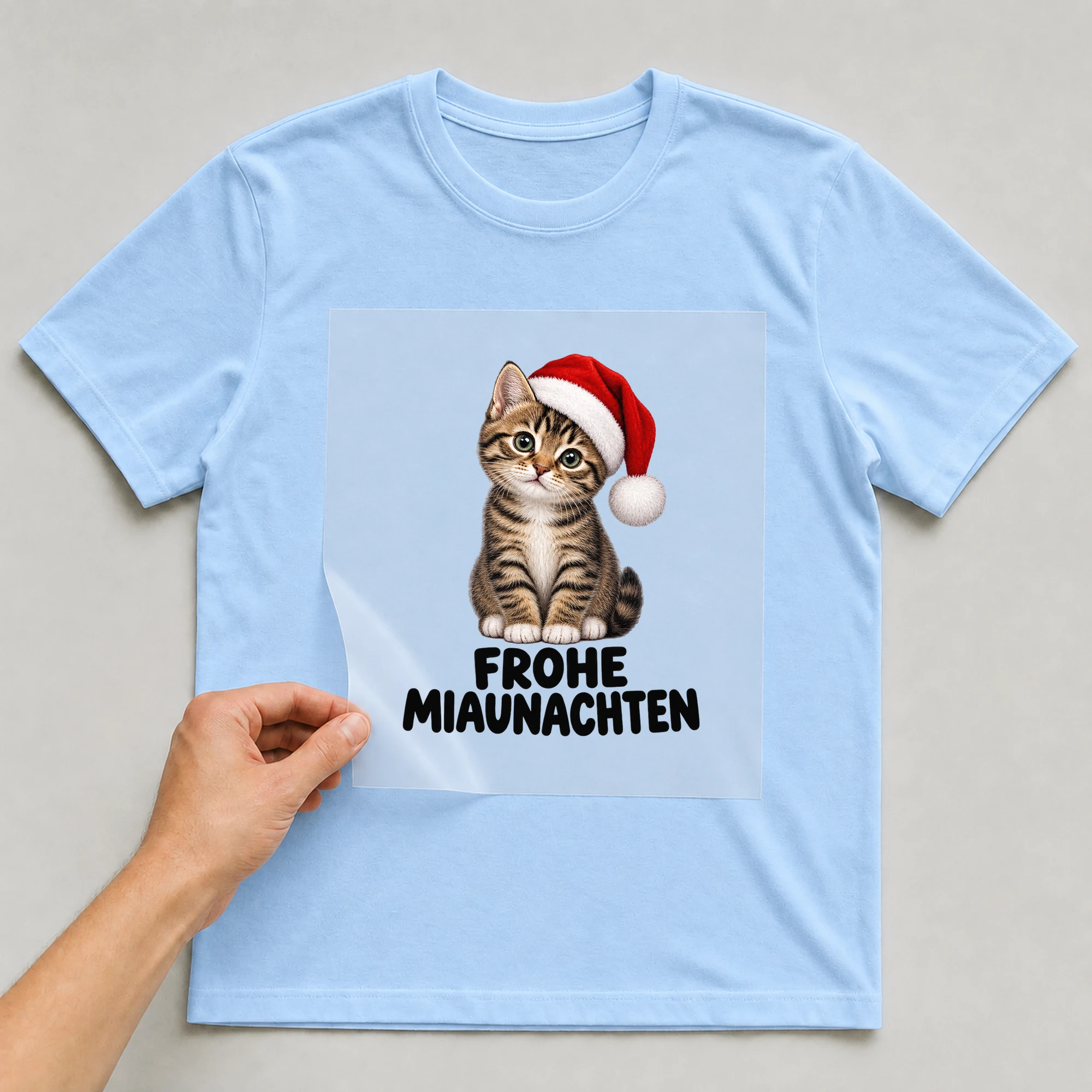 Bügelbild „Weihnachtskatze mit Nikolausmütze“ auf babyblauem T-Shirt, Folie wird aufgelegt