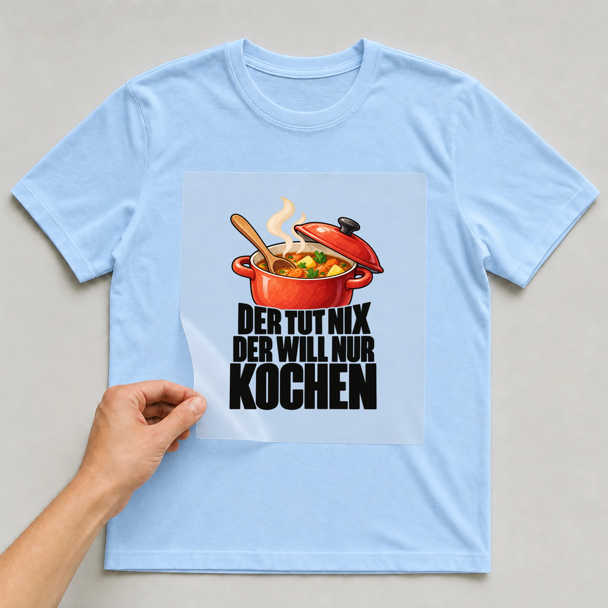 Bügelbild „Der tut nix der will nur kochen“ auf babyblauem T-Shirt, Folie wird aufgelegt