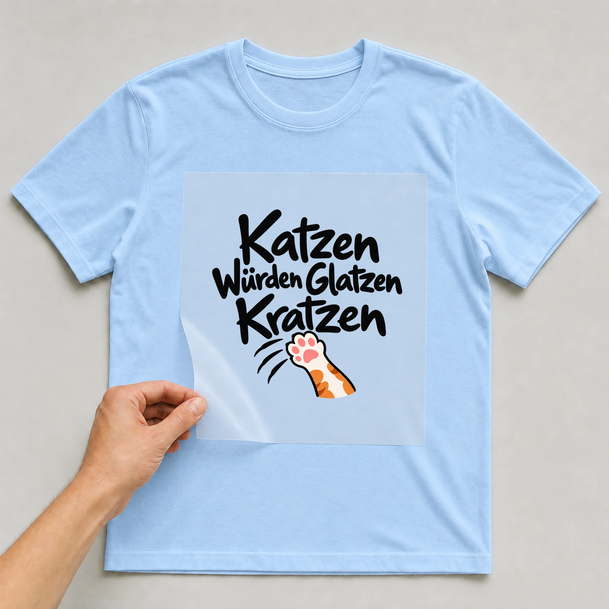 Bügelbild „Katzen Würden Glatzen Kratzen“ auf babyblauem T-Shirt, Folie wird aufgelegt