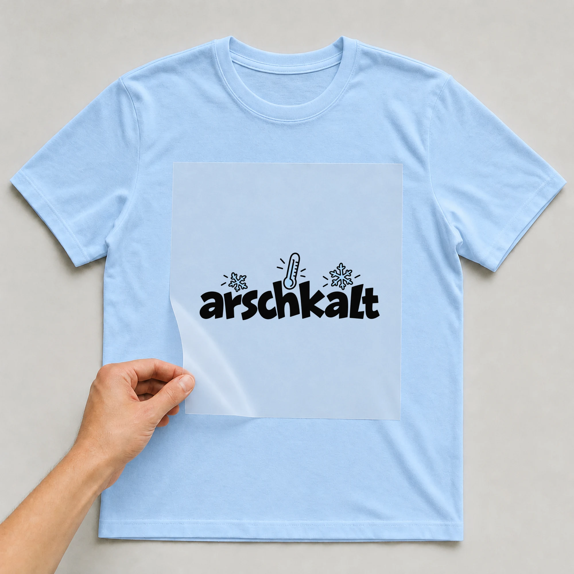 Bügelbild „ar*chkalt“ auf babyblauem T-Shirt, Folie wird aufgelegt