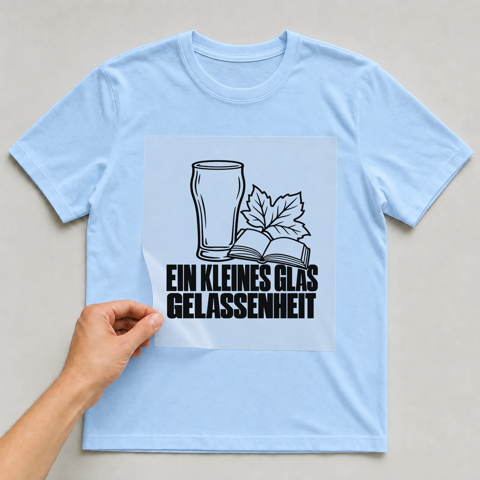 Bügelbild „Ein Kleines Glas Gelassenheit“ auf hellblauem T-Shirt, Folie wird aufgelegt