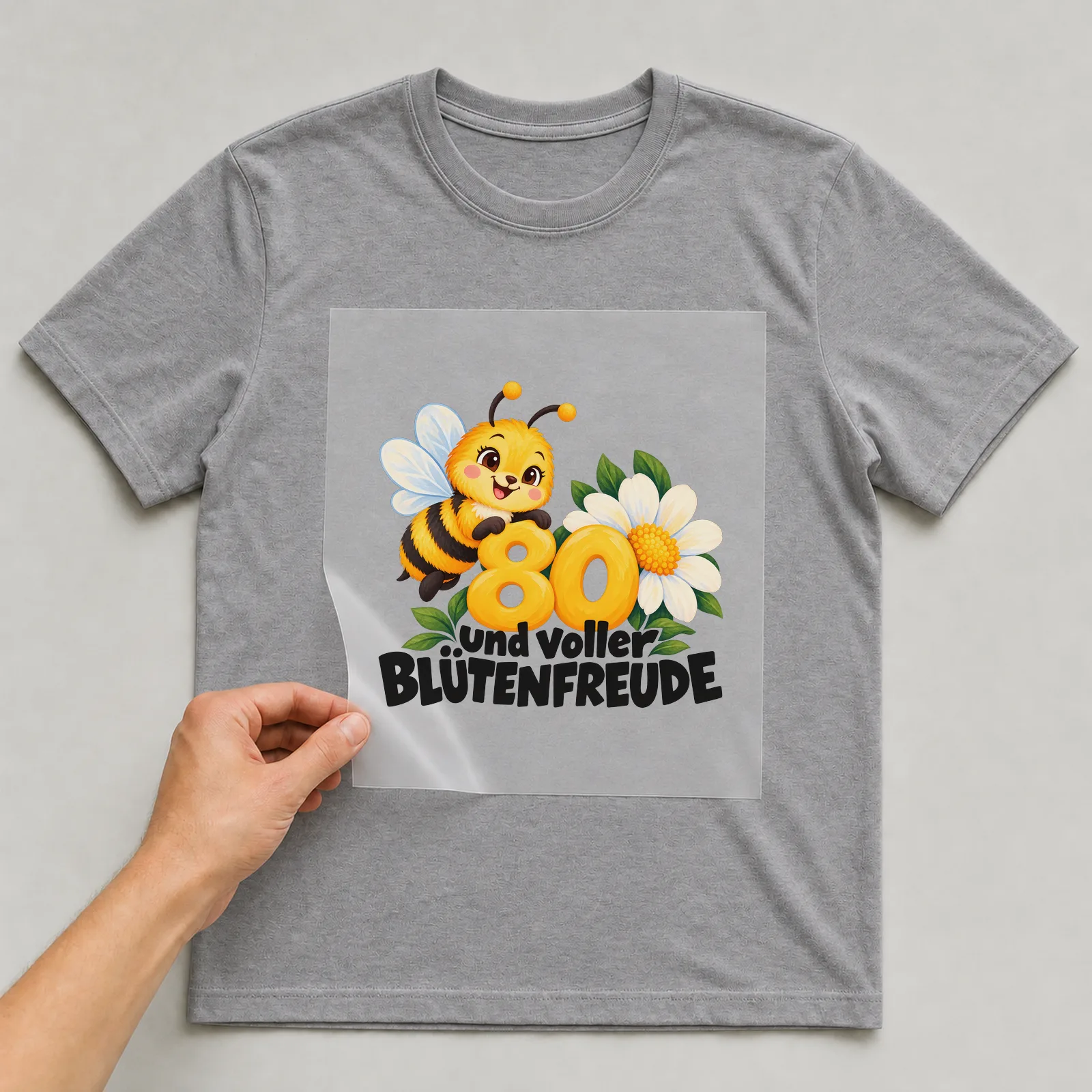 Bügelbild „80 und voller BLÜTENFREUDE“ auf grauem T-Shirt, Folie wird aufgelegt