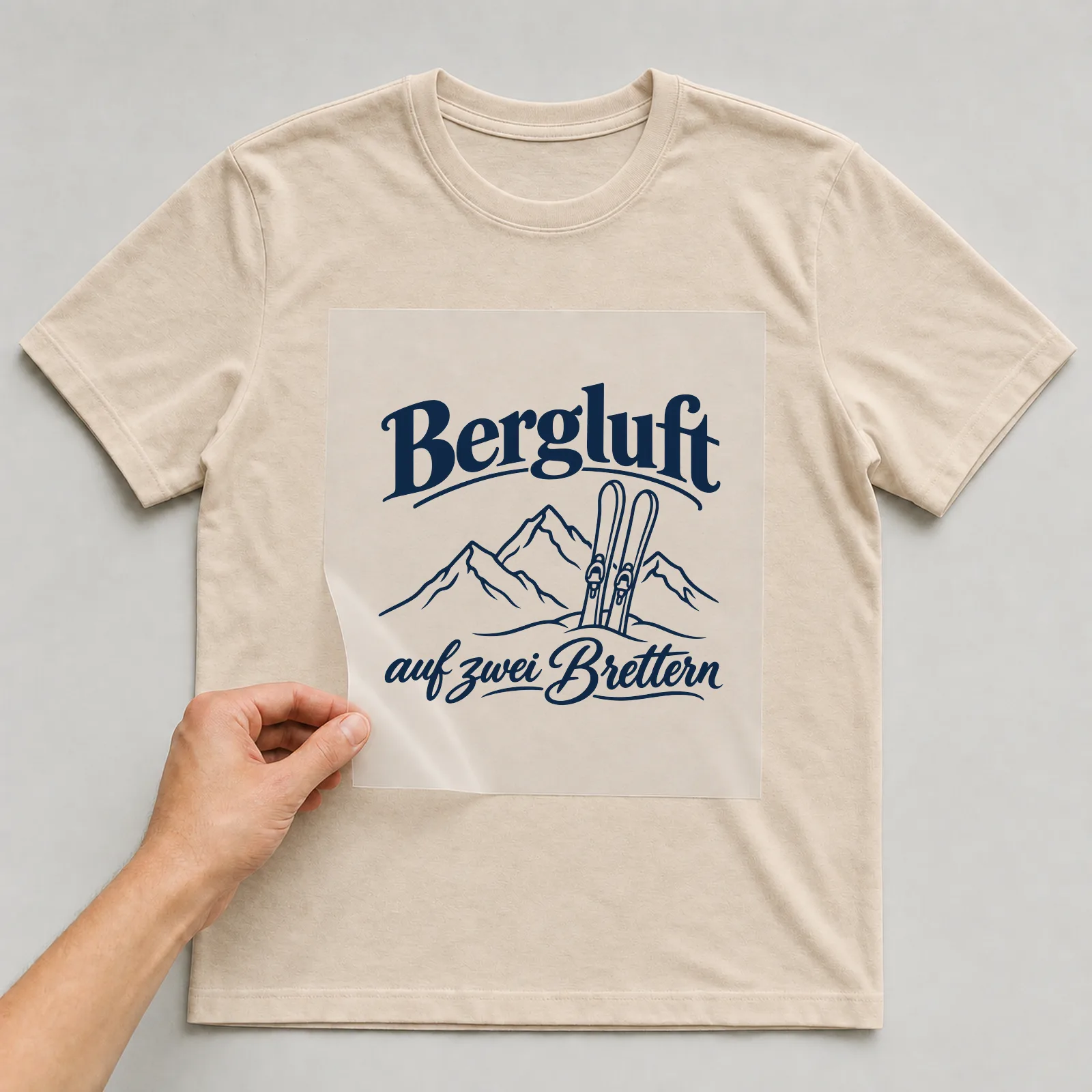 Bügelbild „Bergluft auf zwei Brettern“ auf beigem T-Shirt, Folie wird aufgelegt