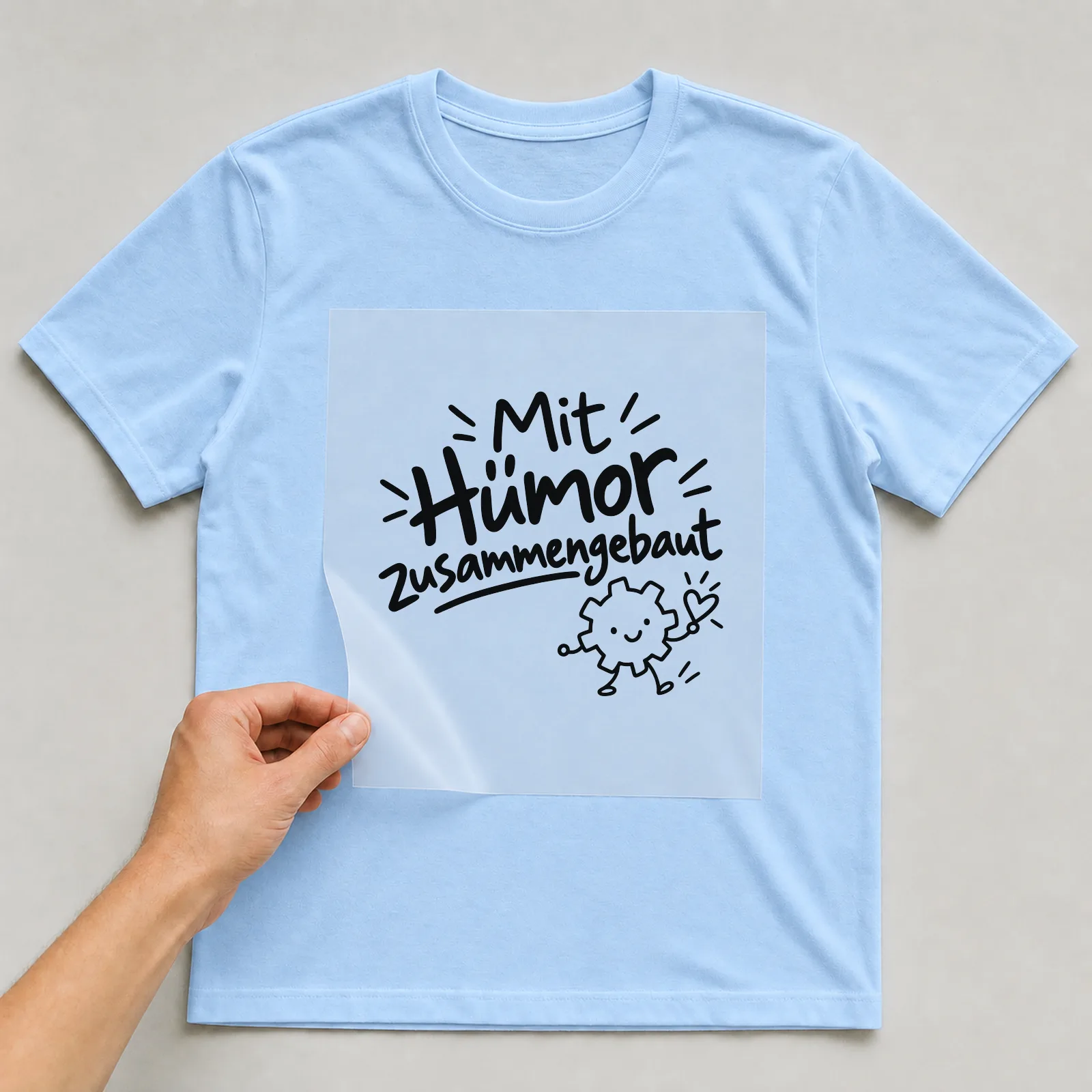 Bügelbild „Mit Humor zusammengebaut“ auf hellblauem T-Shirt, Folie wird aufgelegt