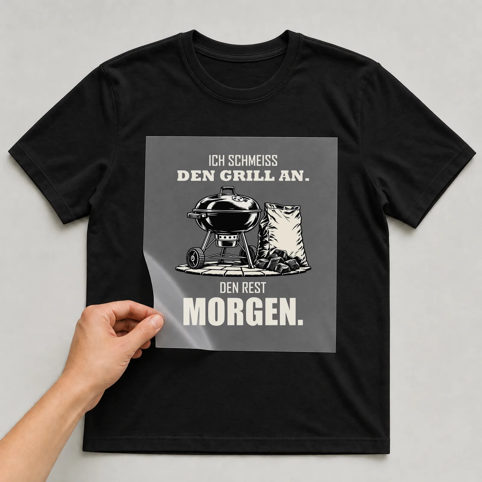 Bügelbild „Ich schmeiß den Grill an. Den Rest morgen.“ auf schwarzem T-Shirt, Folie wird aufgelegt