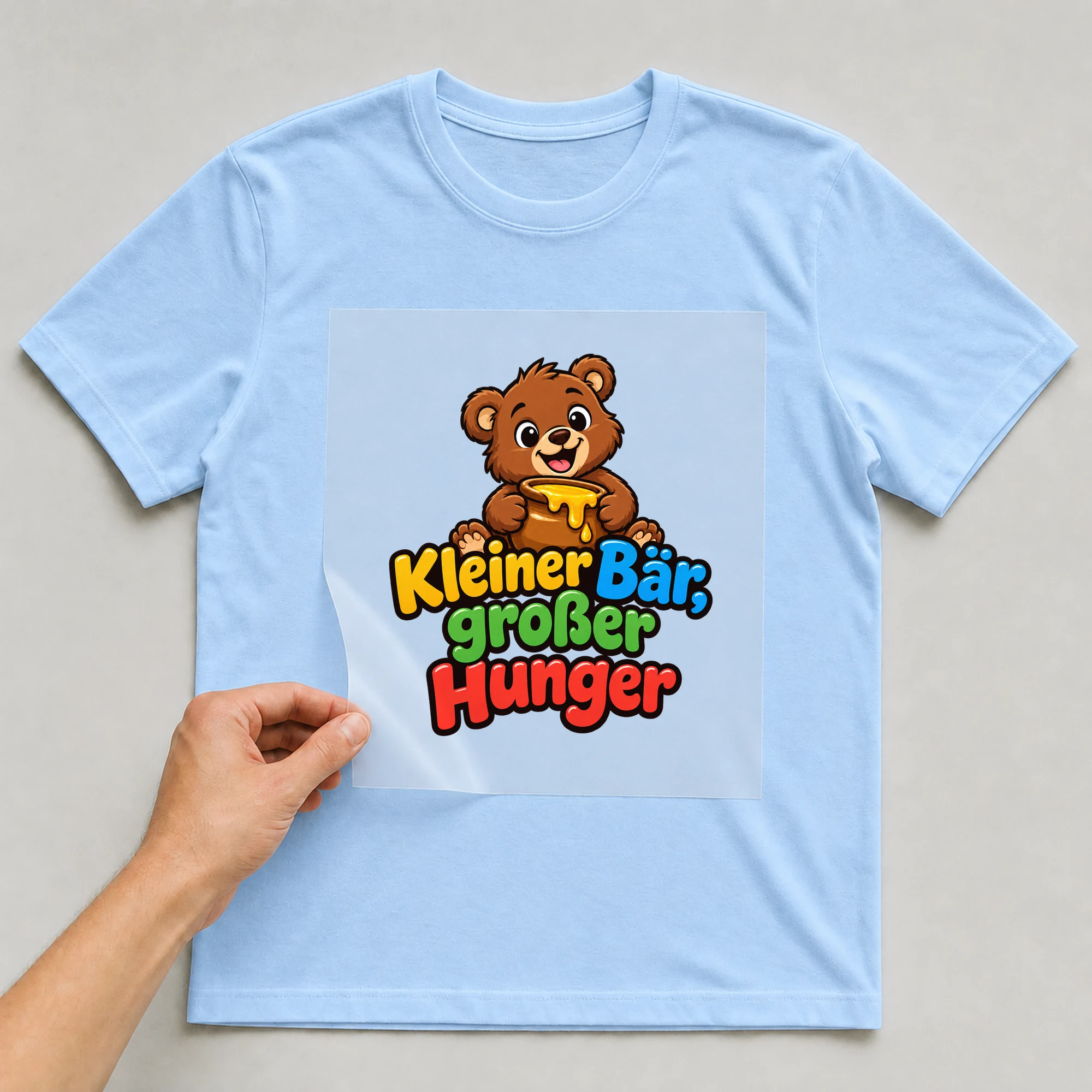 Bügelbild „Kleiner Bär, großer Hunger“ auf babyblauem T-Shirt, Folie wird aufgelegt