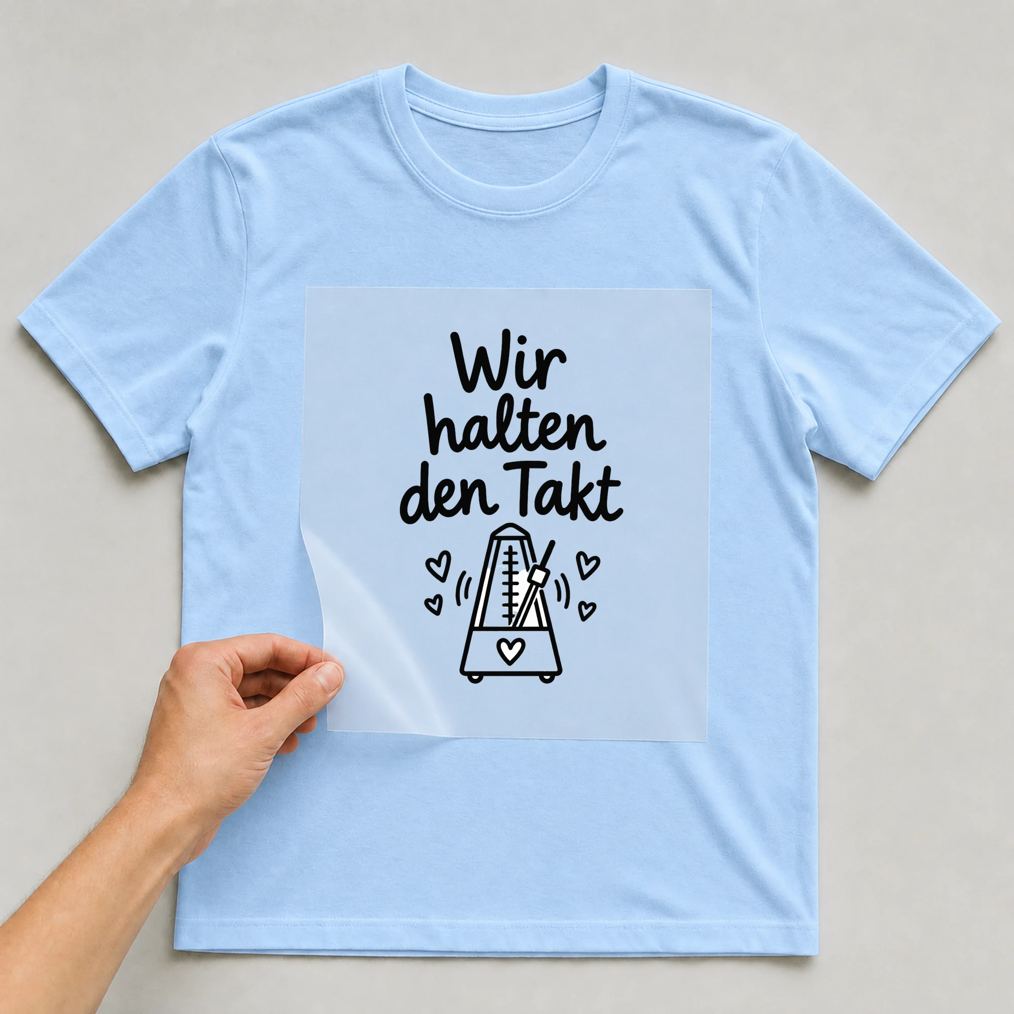 Bügelbild „Wir halten den Takt“ auf babyblauem T-Shirt, Folie wird aufgelegt