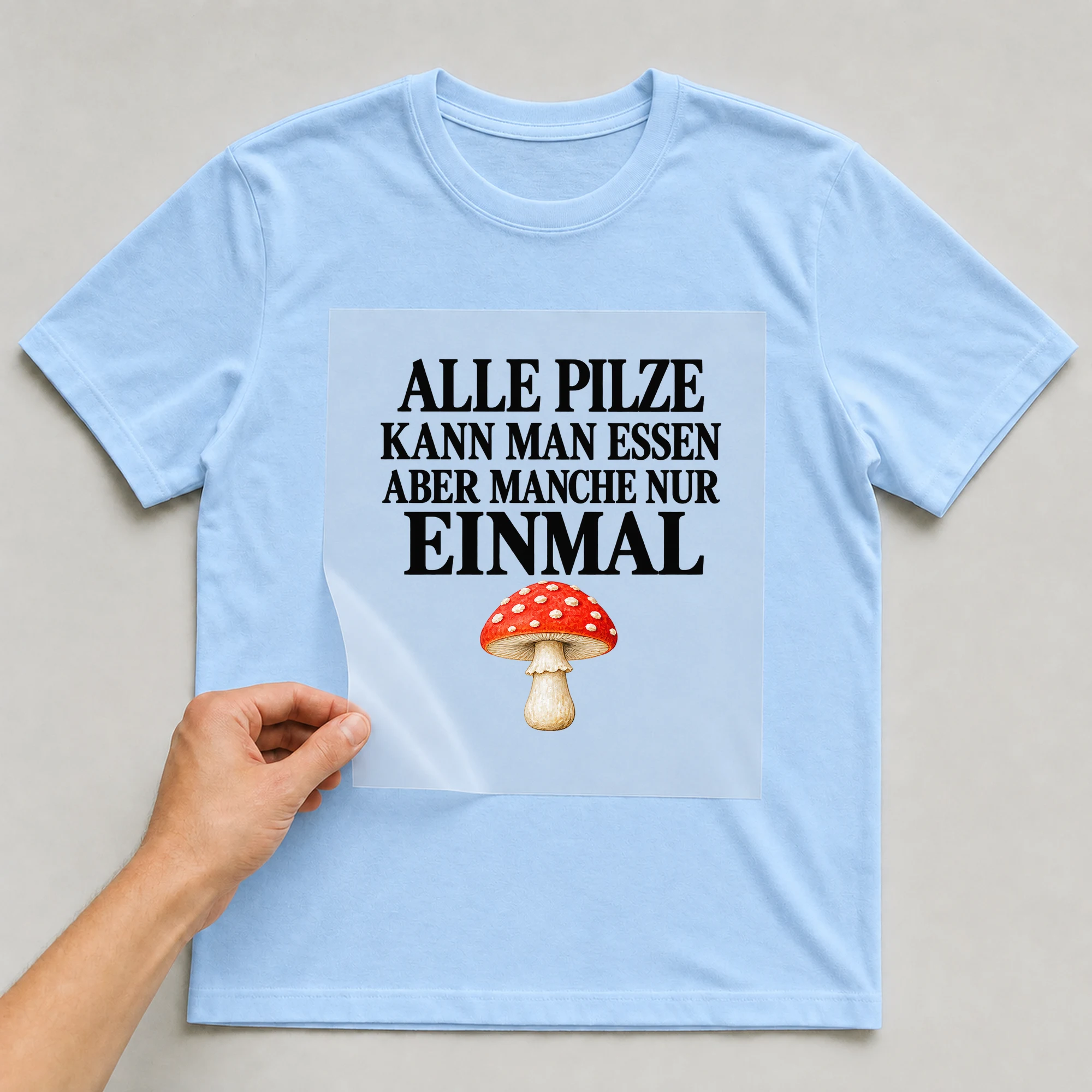 Bügelbild „Alle Pilze kann man Essen aber manche nur einmal“ auf babyblauem T-Shirt, Folie wird aufgelegt