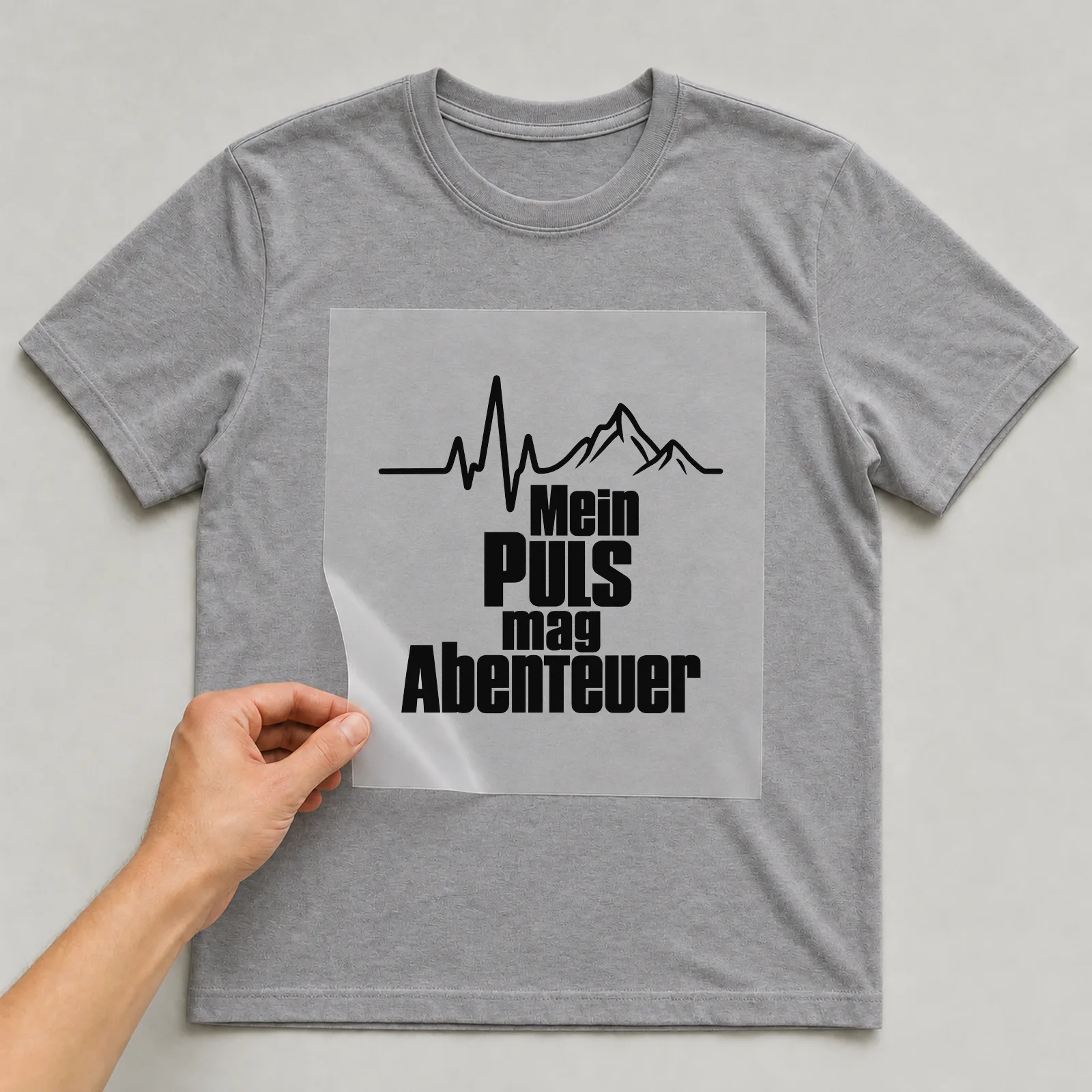 Bügelbild „Mein Puls mag Abenteuer“ auf grauem T-Shirt, Folie wird aufgelegt