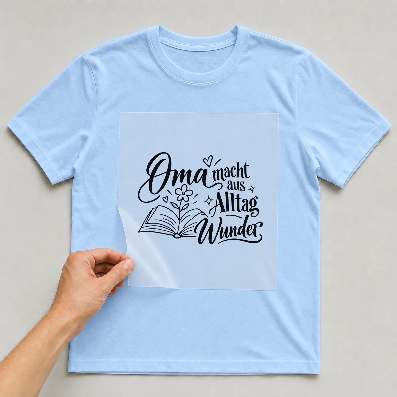 Bügelbild „Oma macht aus Alltag Wunder“ auf hellblauem T-Shirt, Folie wird aufgelegt