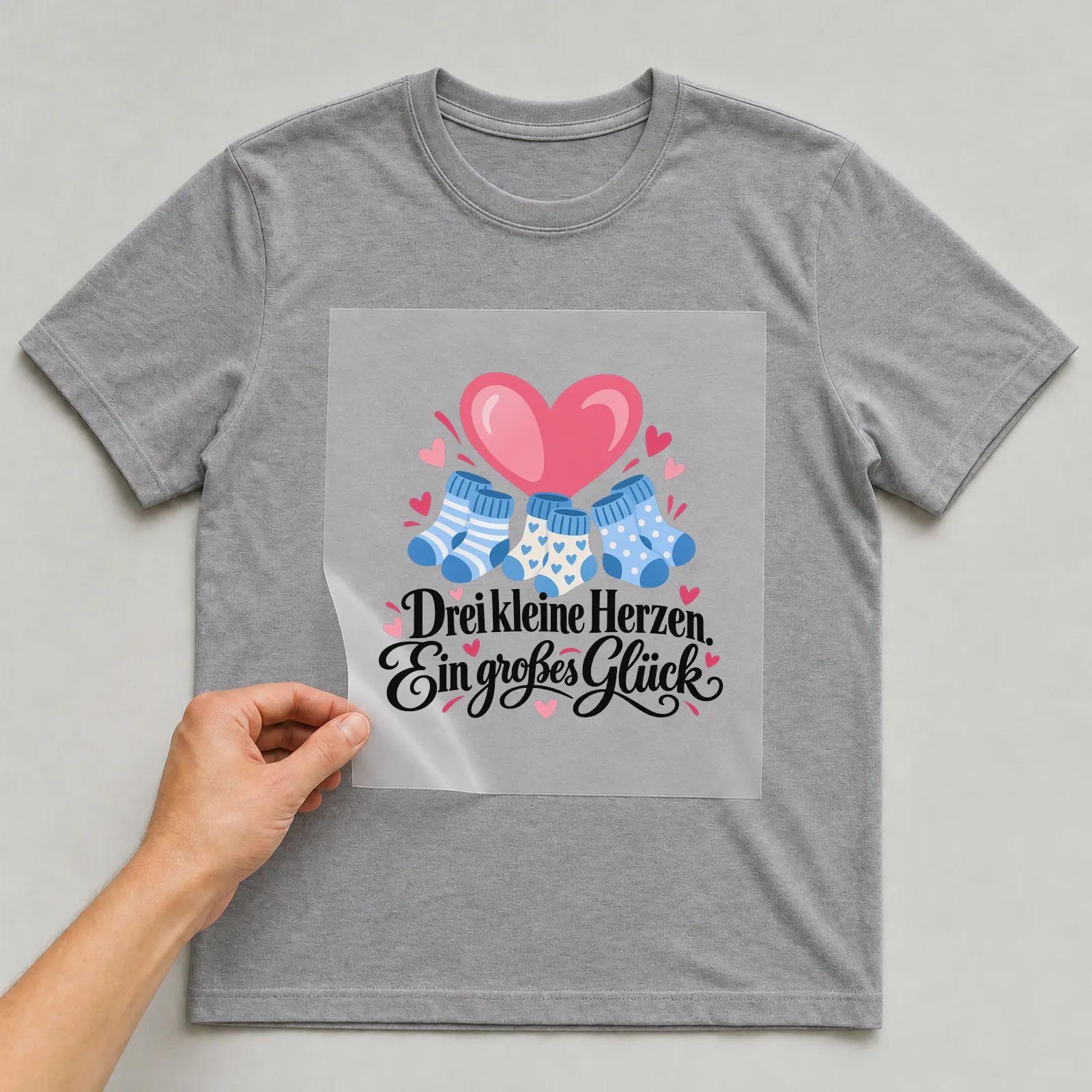 Bügelbild „Drei kleine Herzen. Ein großes Glück“ auf grauem T-Shirt, Folie wird aufgelegt