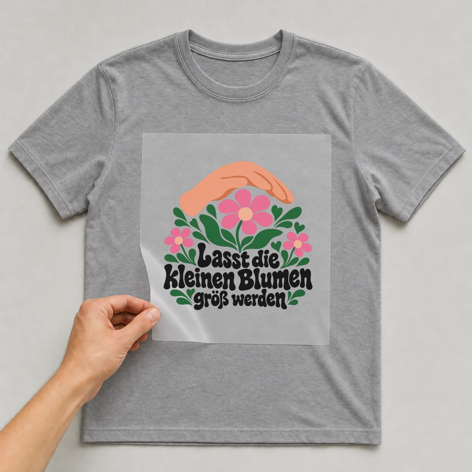 Bügelbild „Lasst die kleinen Blumen groß werden“ auf grauem T-Shirt, Folie wird aufgelegt