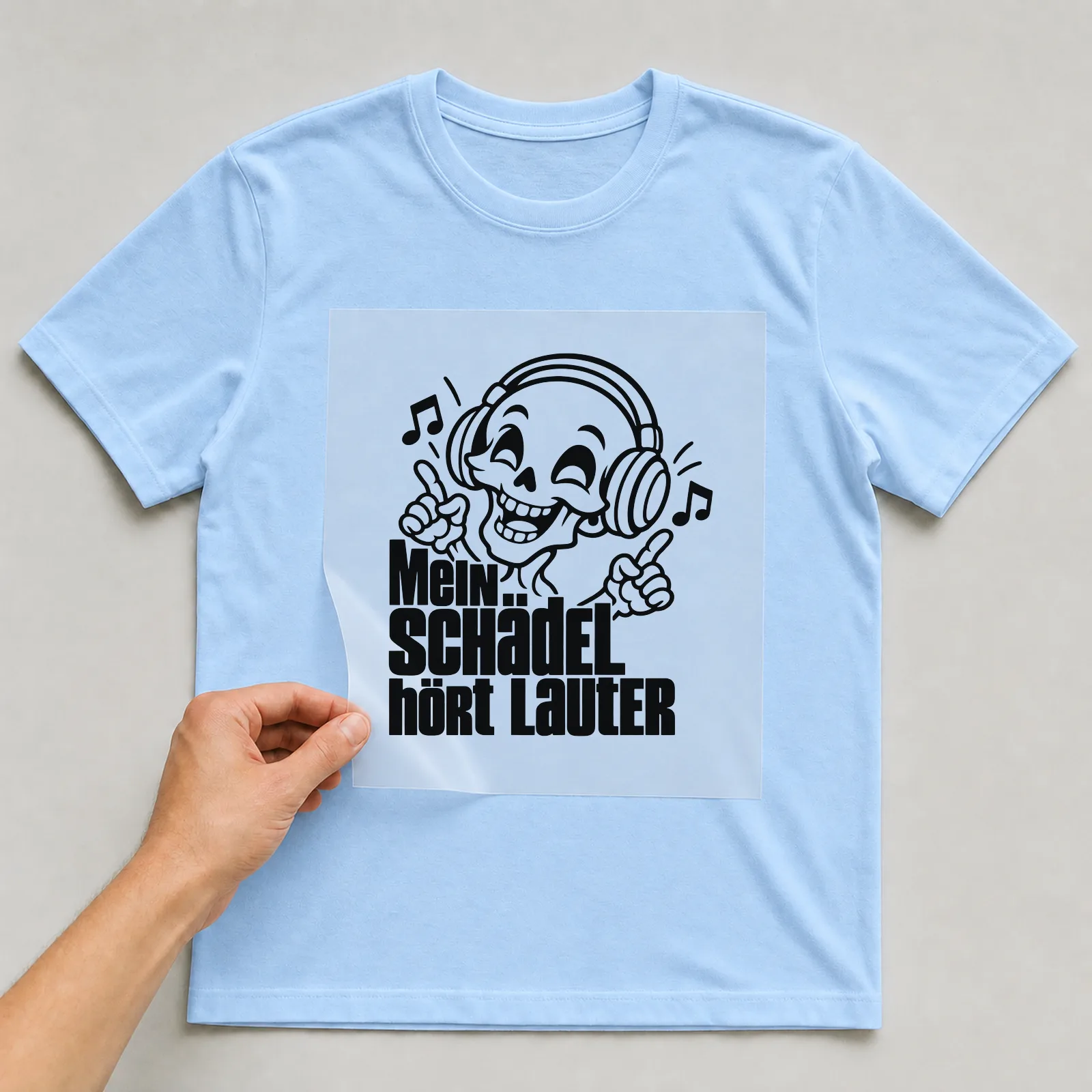 Bügelbild „Mein Schädel hört lauter“ auf hellblauem T-Shirt, Folie wird aufgelegt
