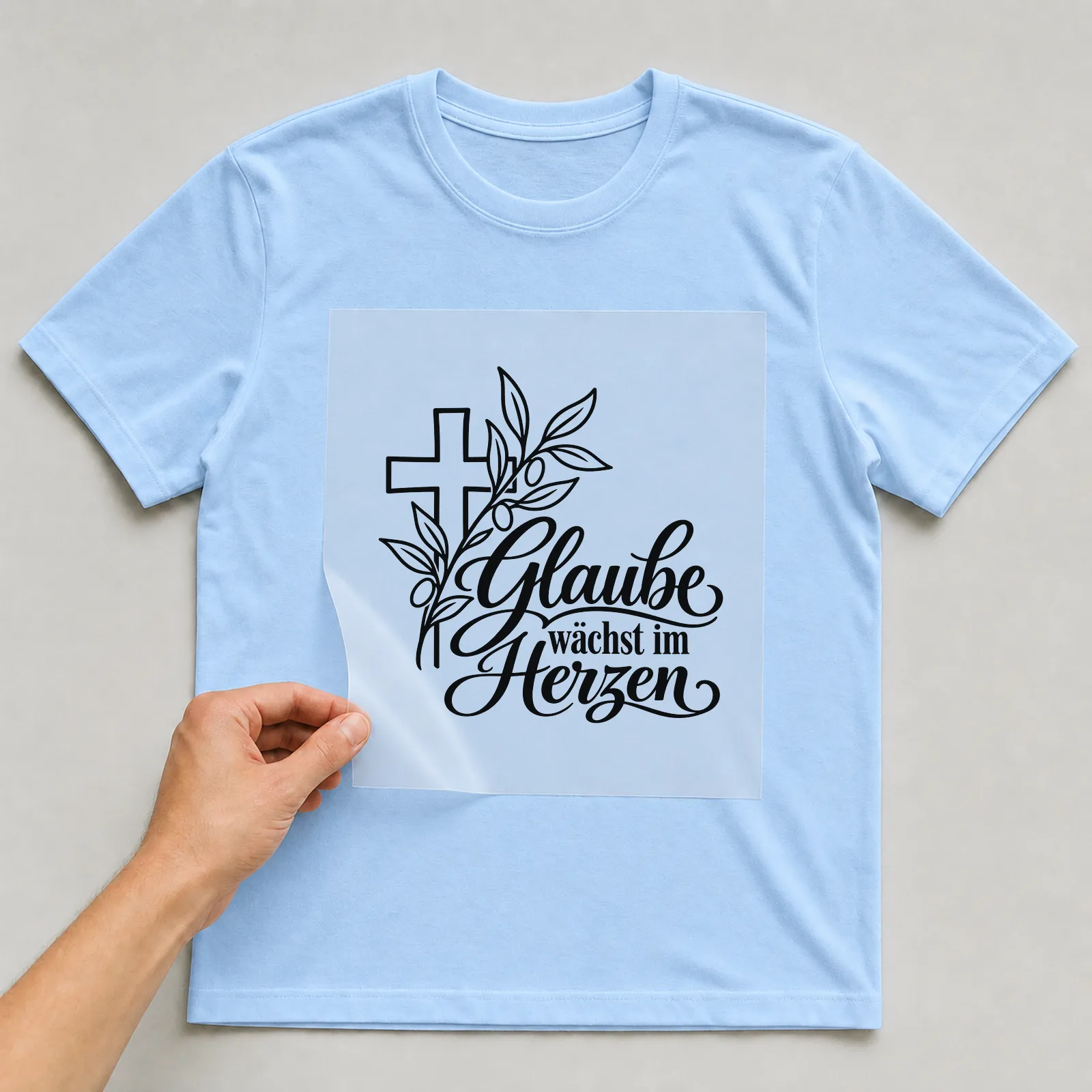 Bügelbild „Glaube wächst im Herzen“ auf hellblauem T-Shirt, Folie wird aufgelegt