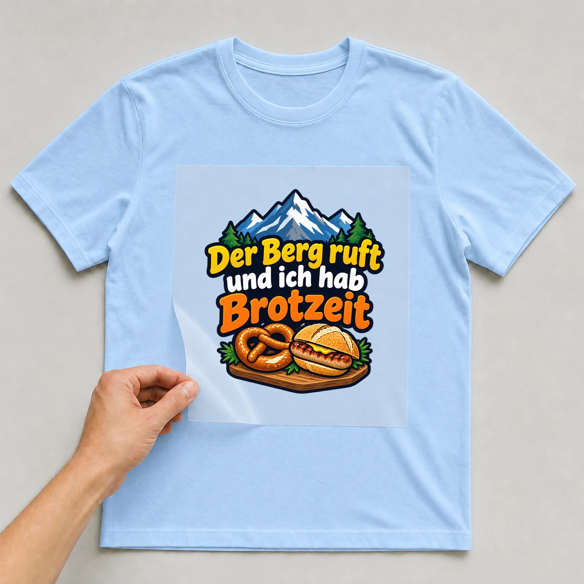 Bügelbild „Der berg ruft und ich hab“ auf babyblauem T-Shirt, Folie wird aufgelegt