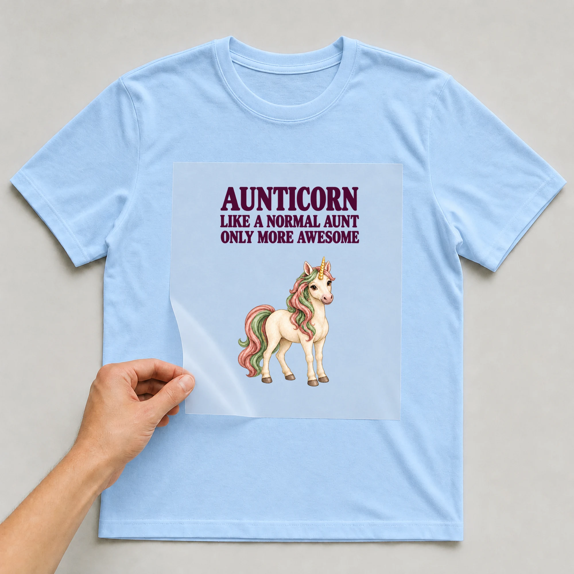 Bügelbild „AUNTICORN LIKE A NORMAL AUNT ONLY MORE AWESOME“ auf babyblauem T-Shirt, Folie wird aufgelegt