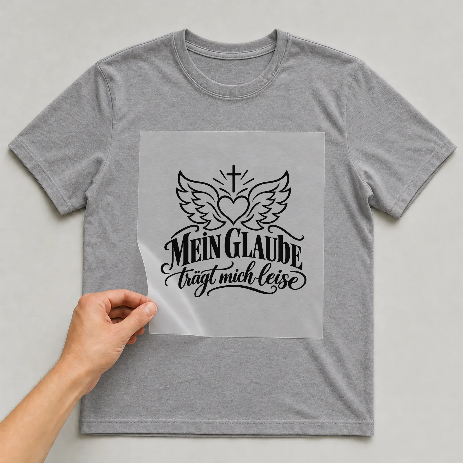 Bügelbild „MEIN GLAUBE trägt mich leise“ auf grauem T-Shirt, Folie wird aufgelegt