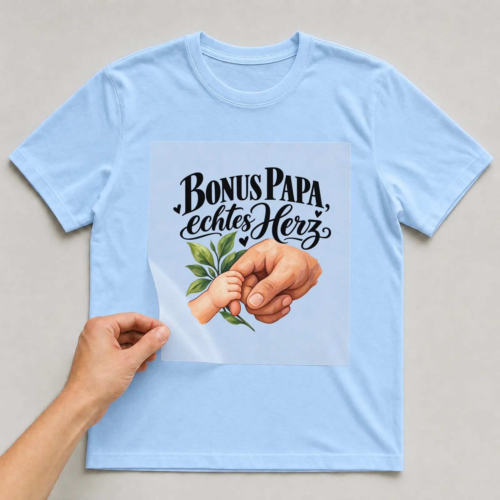 Bügelbild „Bonus Papa, echtes Herz“ auf hellblauem T-Shirt, Folie wird aufgelegt