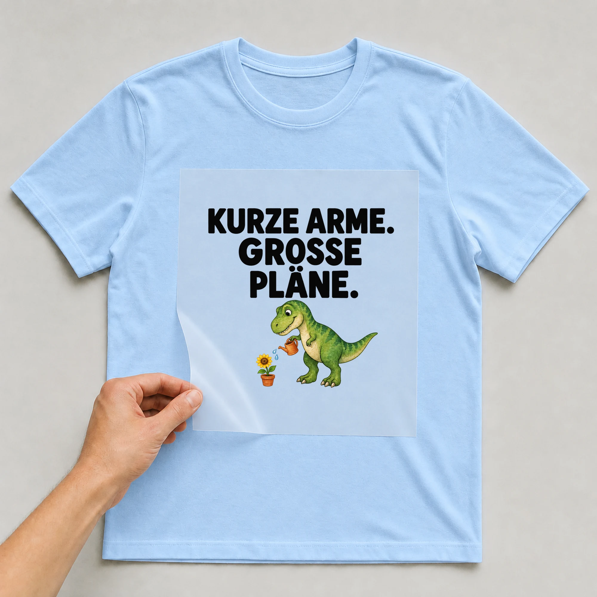 Bügelbild „KURZE ARME. GROSSE PLÄNE.“ auf babyblauem T-Shirt, Folie wird aufgelegt