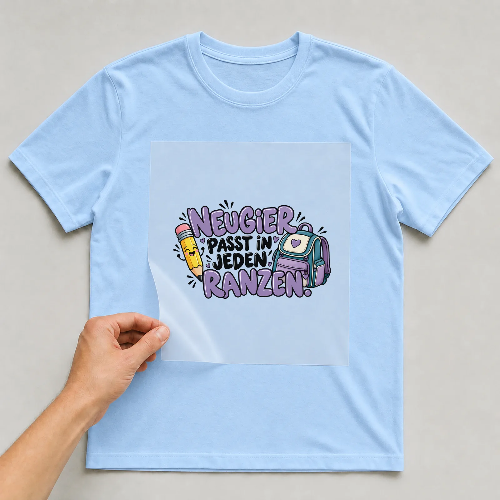 Bügelbild „Neugier passt in jeden Ranzen“ auf hellblauem T-Shirt, Folie wird aufgelegt