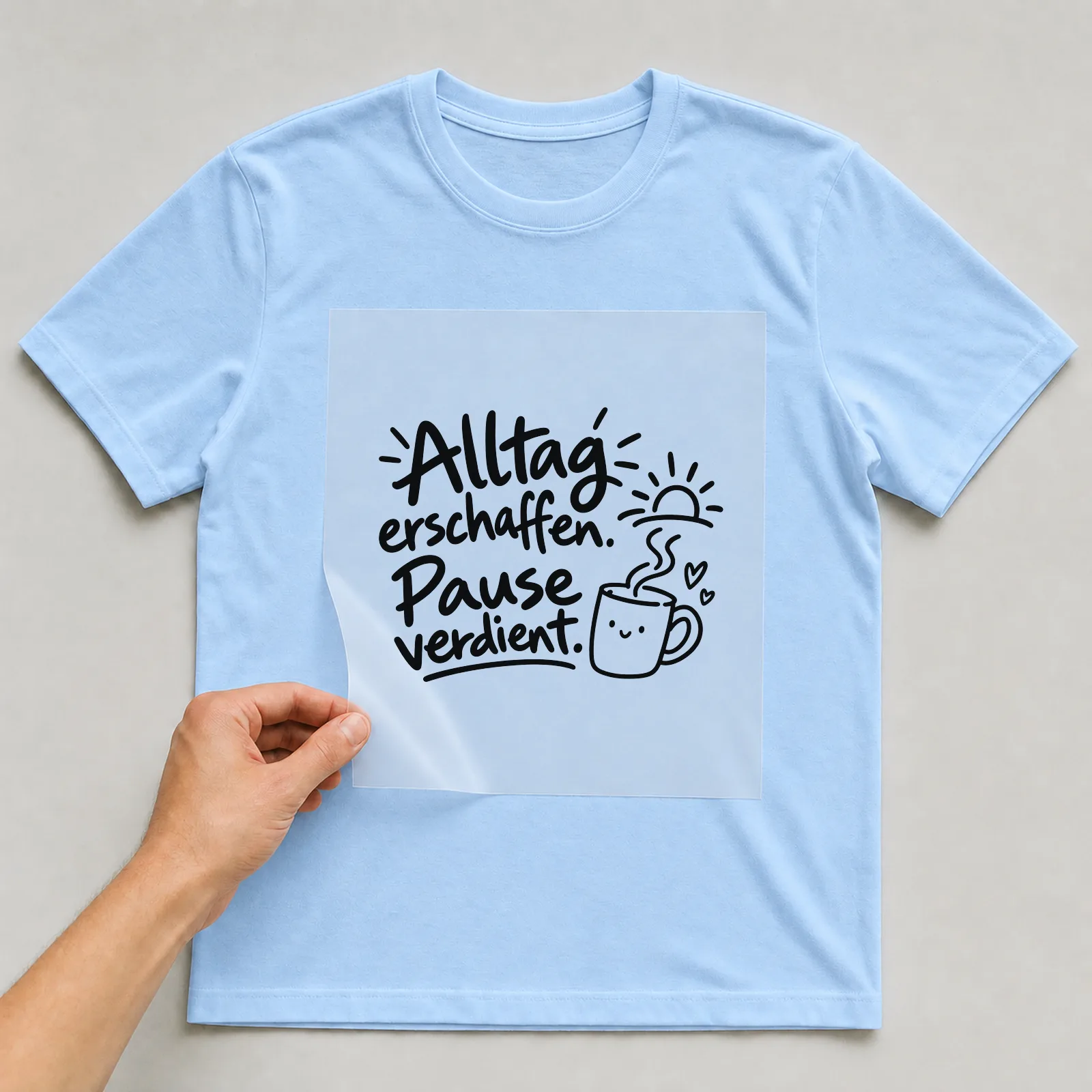 Bügelbild „Alltag erschaffen. Pause verdient.“ auf hellblauem T-Shirt, Folie wird aufgelegt
