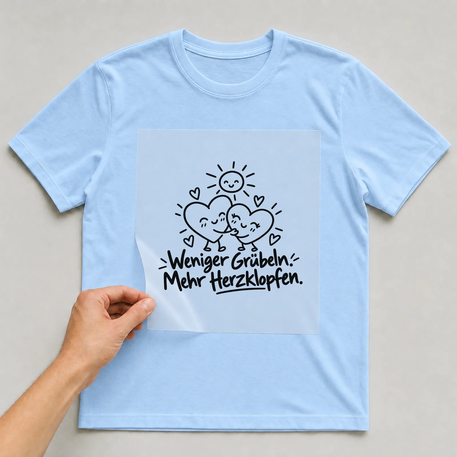 Bügelbild „Weniger Grübeln. Mehr Herzklopfen.“ auf hellblauem T-Shirt, Folie wird aufgelegt