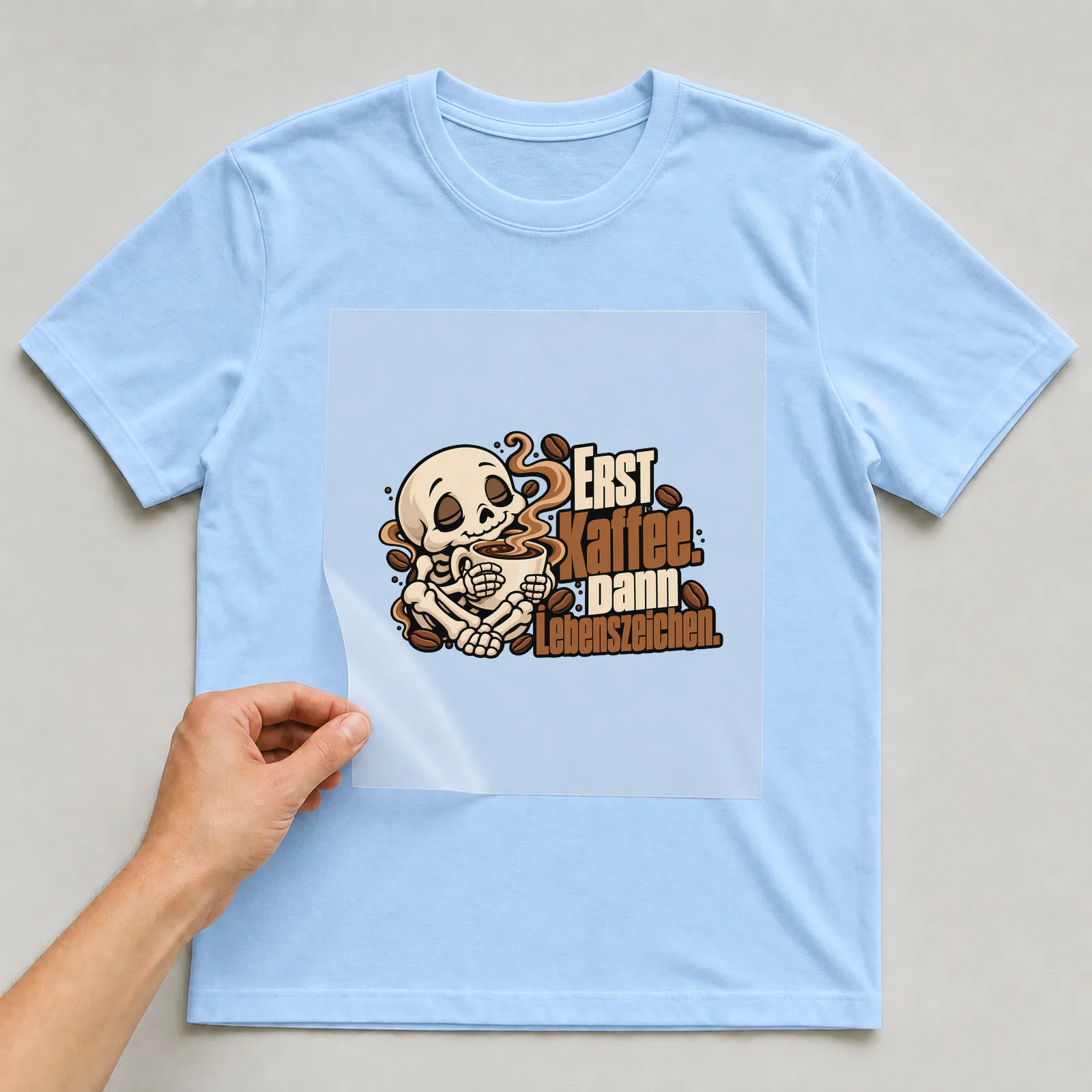 Bügelbild „Erst Kaffee. Dann Lebenszeichen.“ auf hellblauem T-Shirt, Folie wird aufgelegt