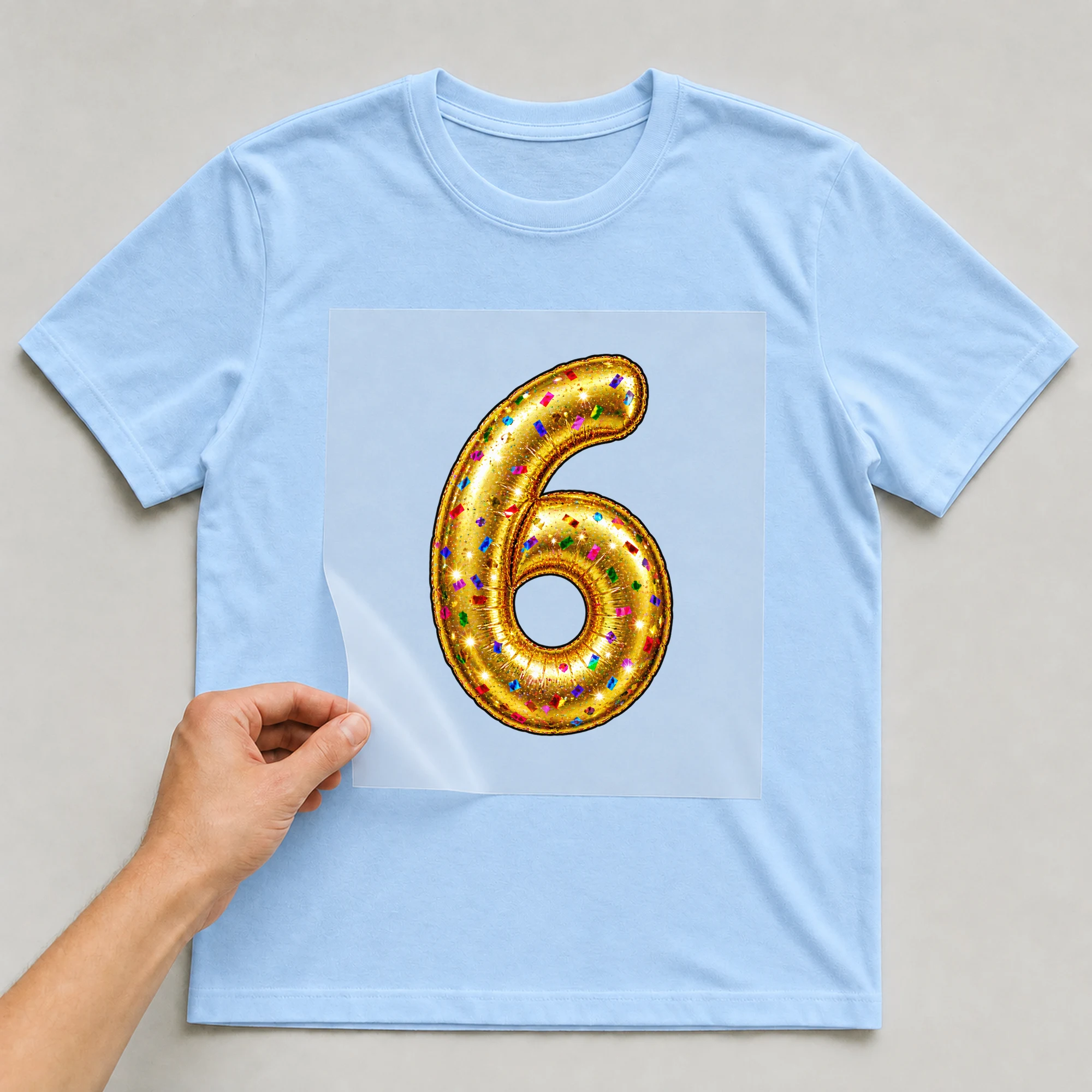 Bügelbild „Zahl 6 im Konfetti-Metallic-Look“ auf babyblauem T-Shirt, Folie wird aufgelegt