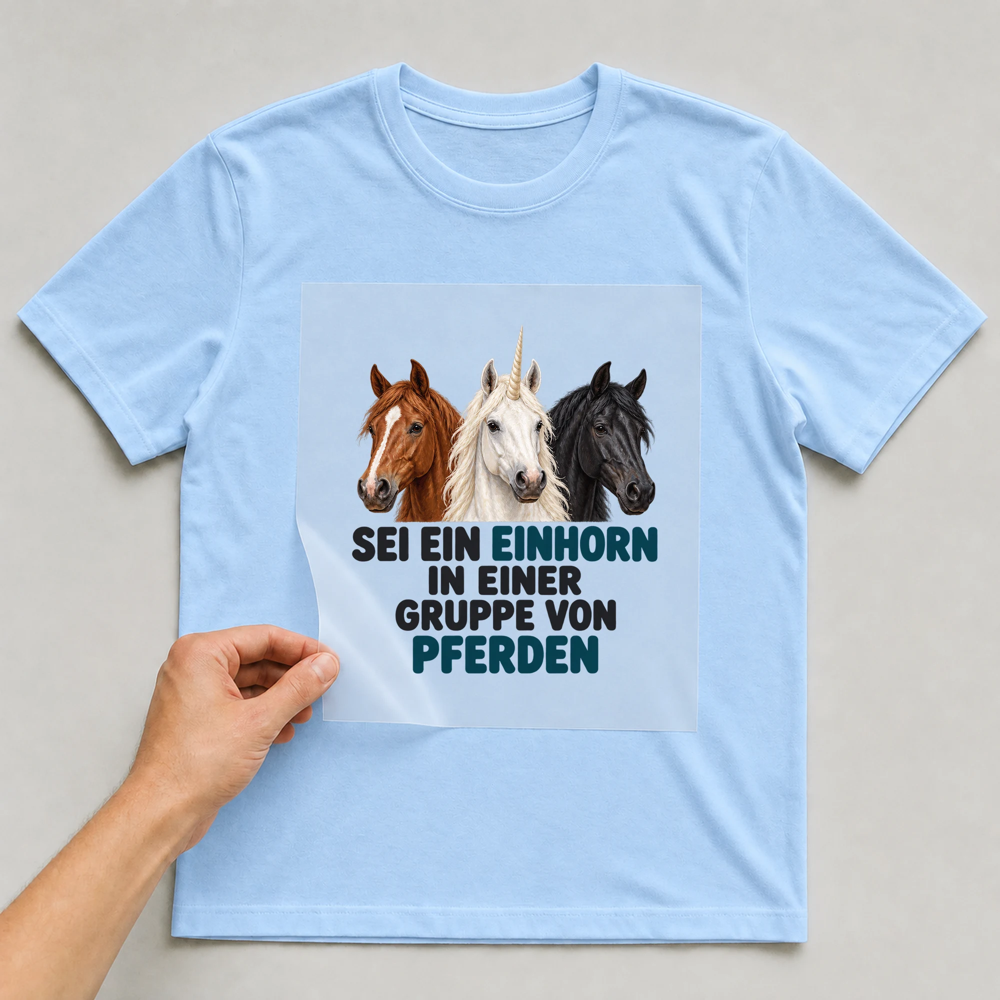 Bügelbild „Sei ein Einhorn in einer Gruppe von pferden“ auf babyblauem T-Shirt, Folie wird aufgelegt
