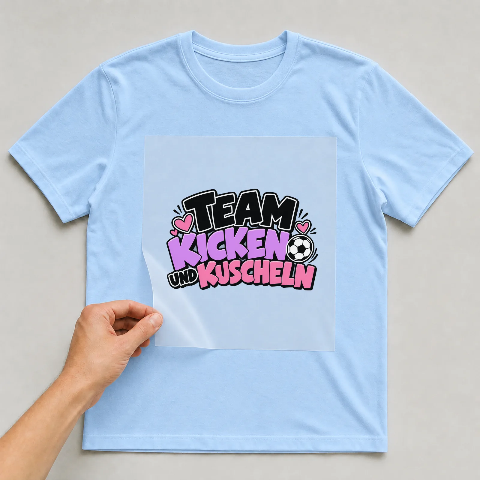 Bügelbild „Team Kicken und Kuscheln“ auf hellblauem T-Shirt, Folie wird aufgelegt