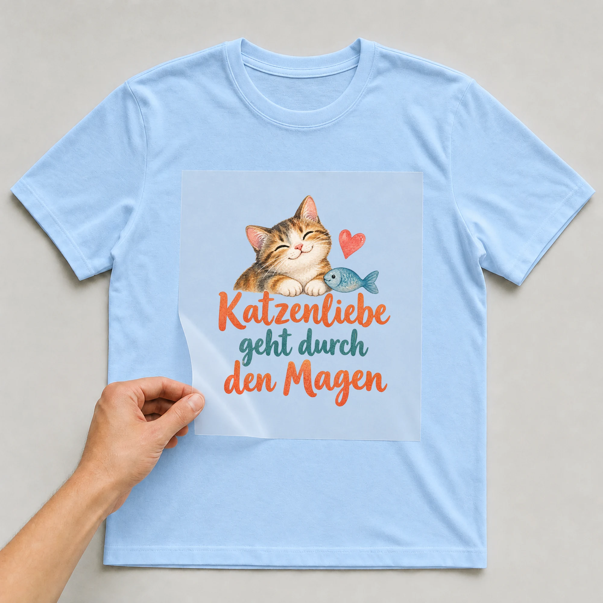 Bügelbild „Katzenliebe geht durch den Magen“ auf babyblauem T-Shirt, Folie wird aufgelegt
